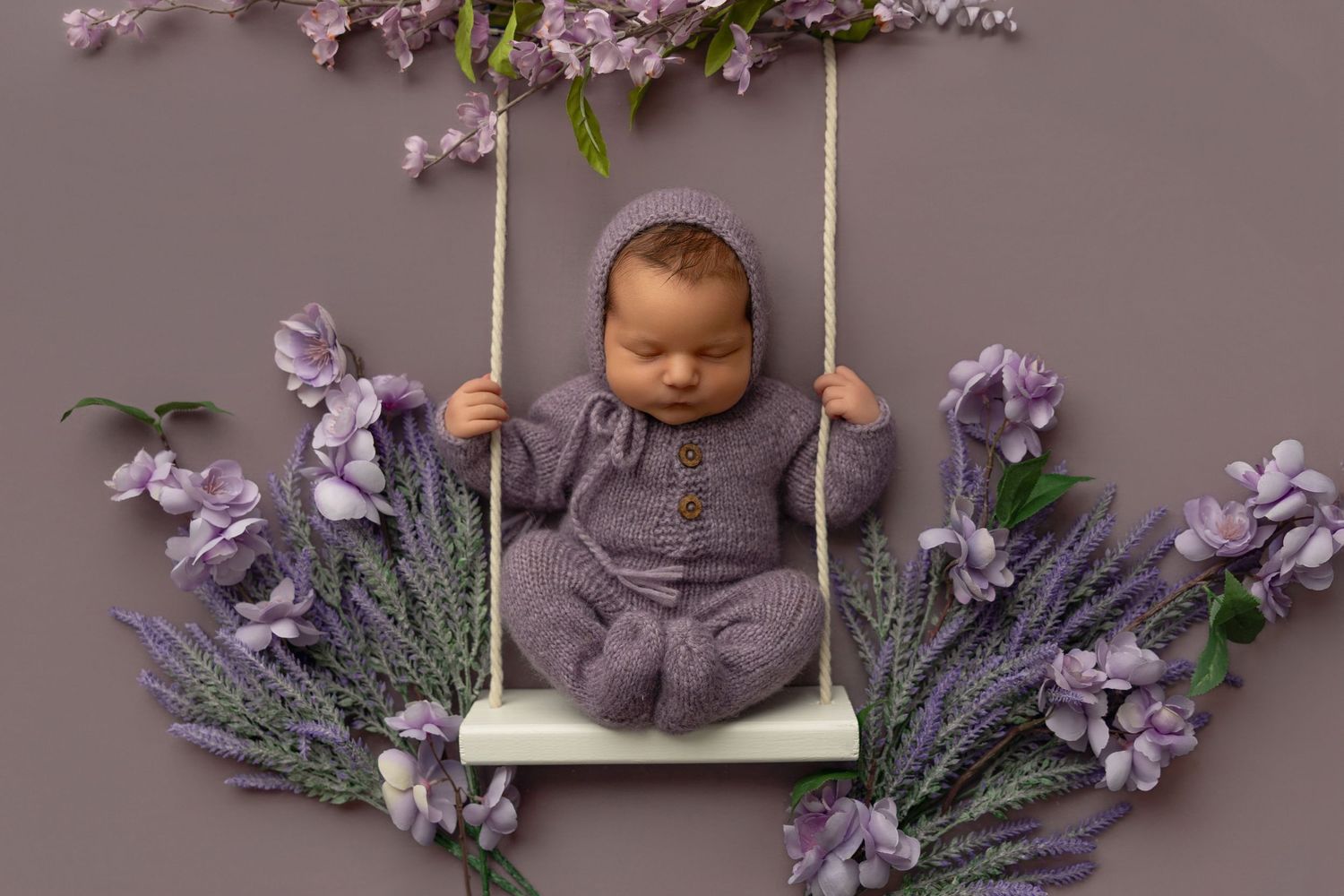 Newborn in gray outfit sits on small white swing surrounded by purple lavender flowers and blossoms.