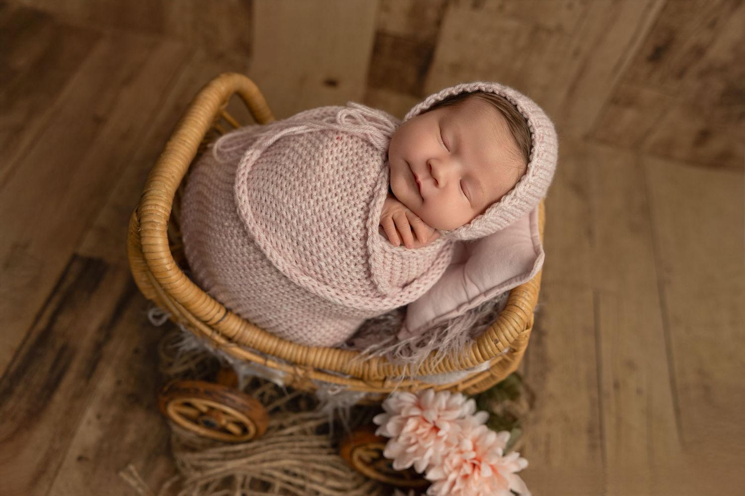 Newborn baby sleeping peacefully in a woven basket on a wooden floor with soft knit blankets and floral accents.