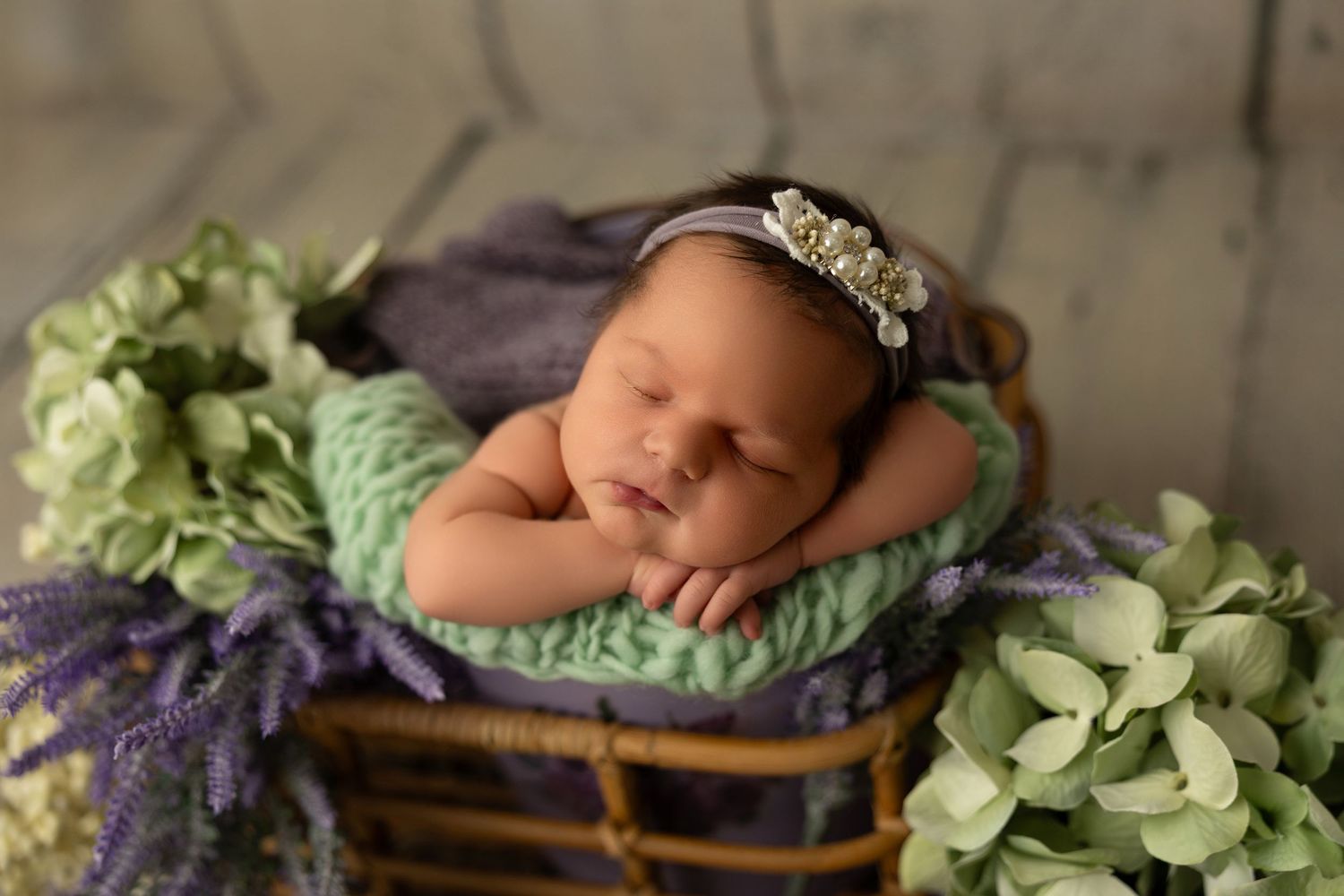 Newborn baby sleeping peacefully in wooden basket surrounded by soft green and purple floral decorations.