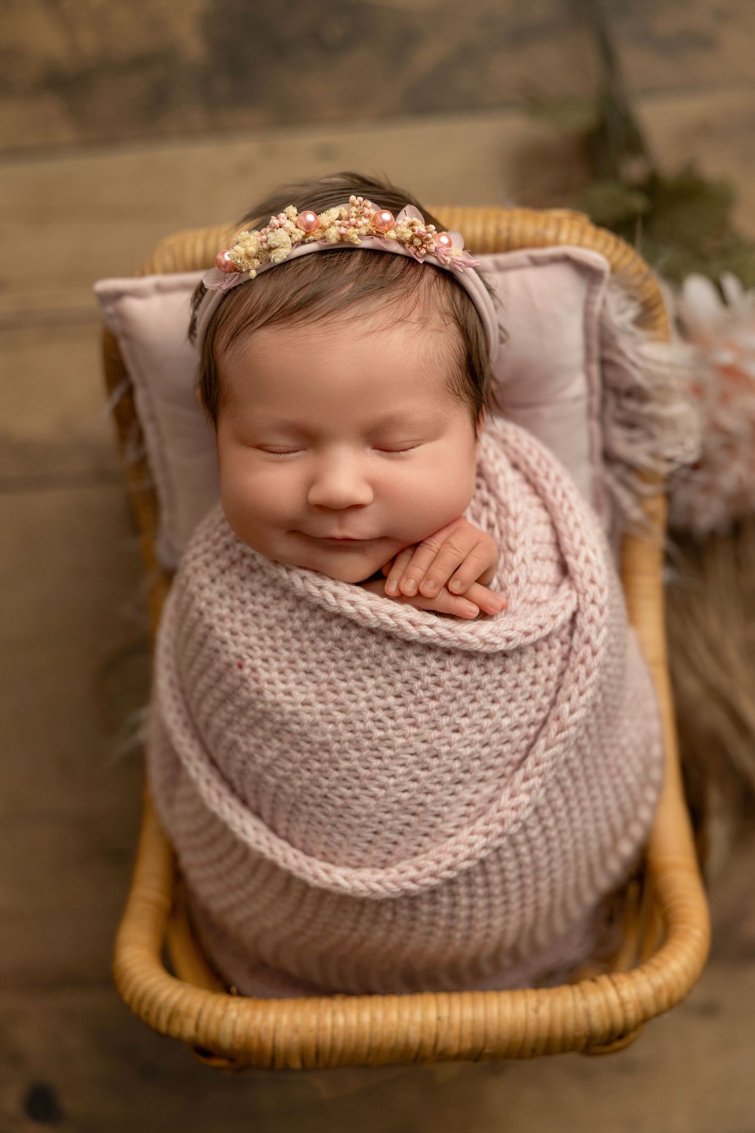 A sleeping newborn wrapped in soft pink knit blanket rests peacefully in a woven basket during professional photography session.