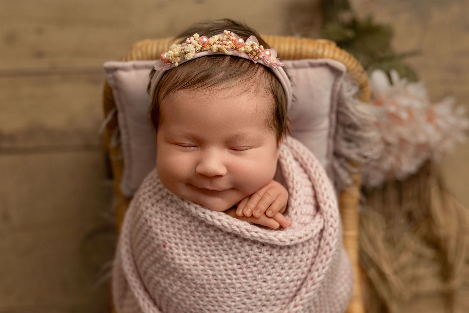 Sleeping baby wrapped in soft pink knit blanket wearing floral headband rests peacefully on cushioned surface.