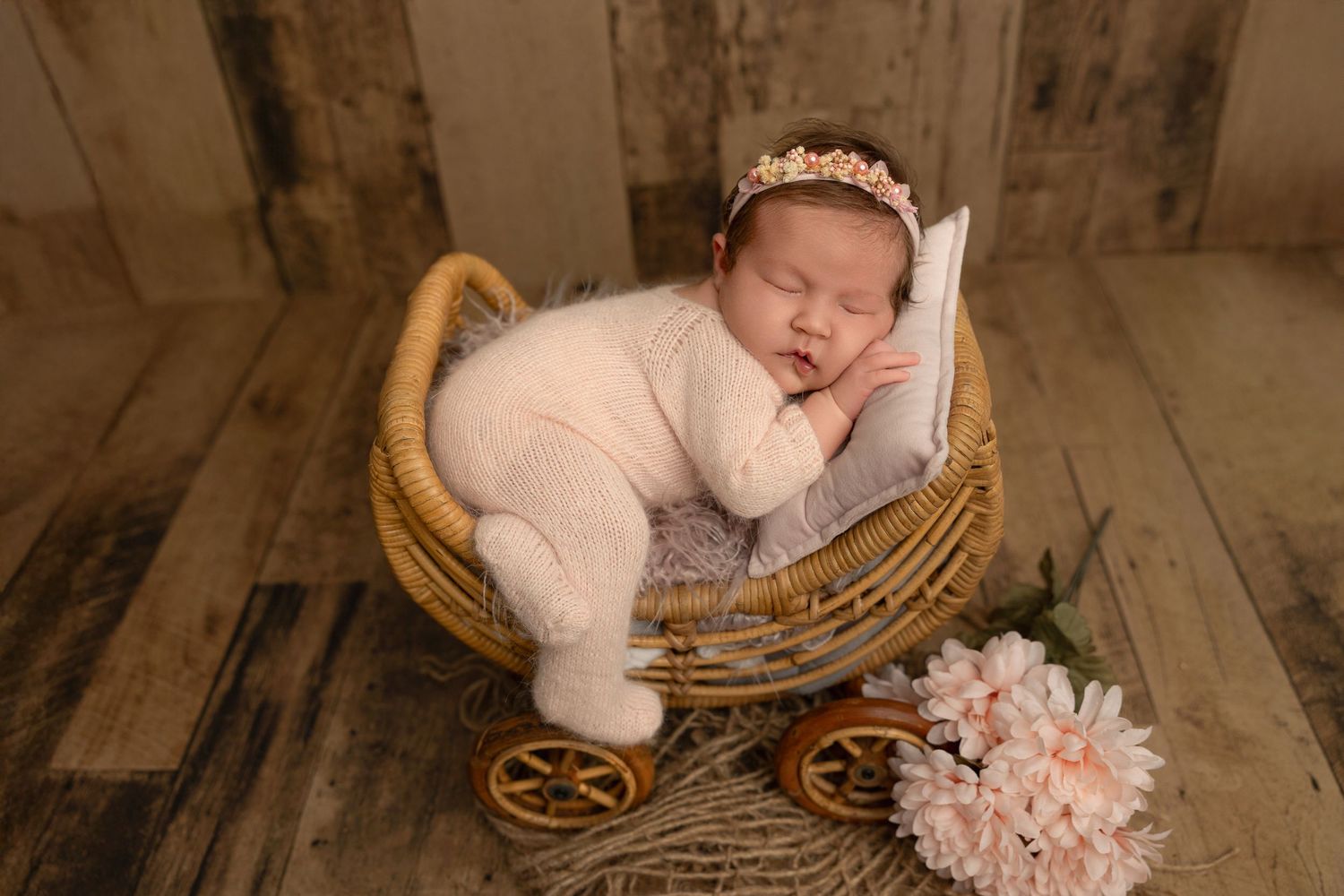 Sleeping newborn in pink outfit rests peacefully in vintage wicker bassinet with floral decoration on wooden floor.