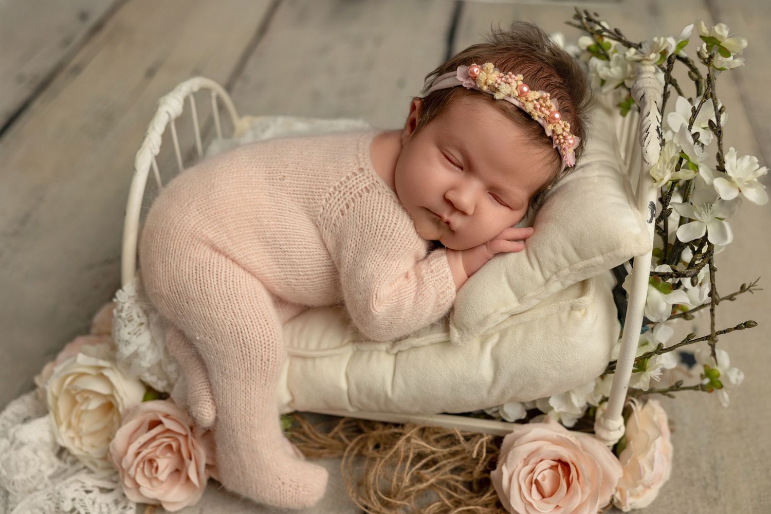 Sleeping newborn baby wrapped in pink swaddle surrounded by delicate white and blush roses and floral decorations.