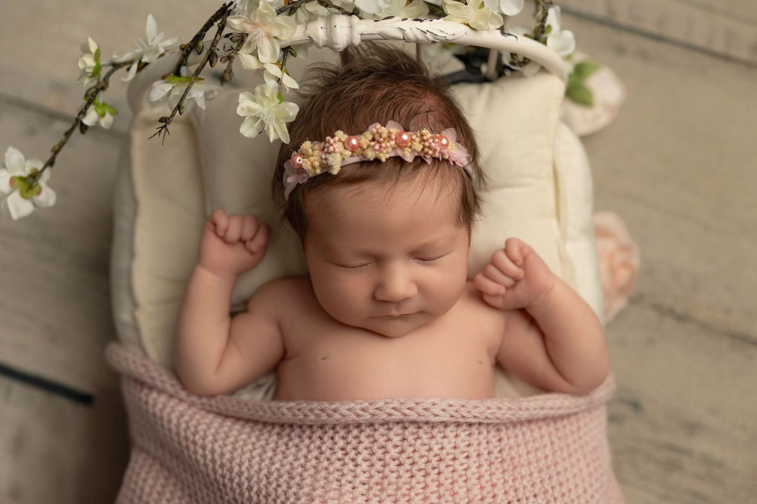 Sleeping baby wrapped in pink blanket with floral headband lying under white cherry blossoms.