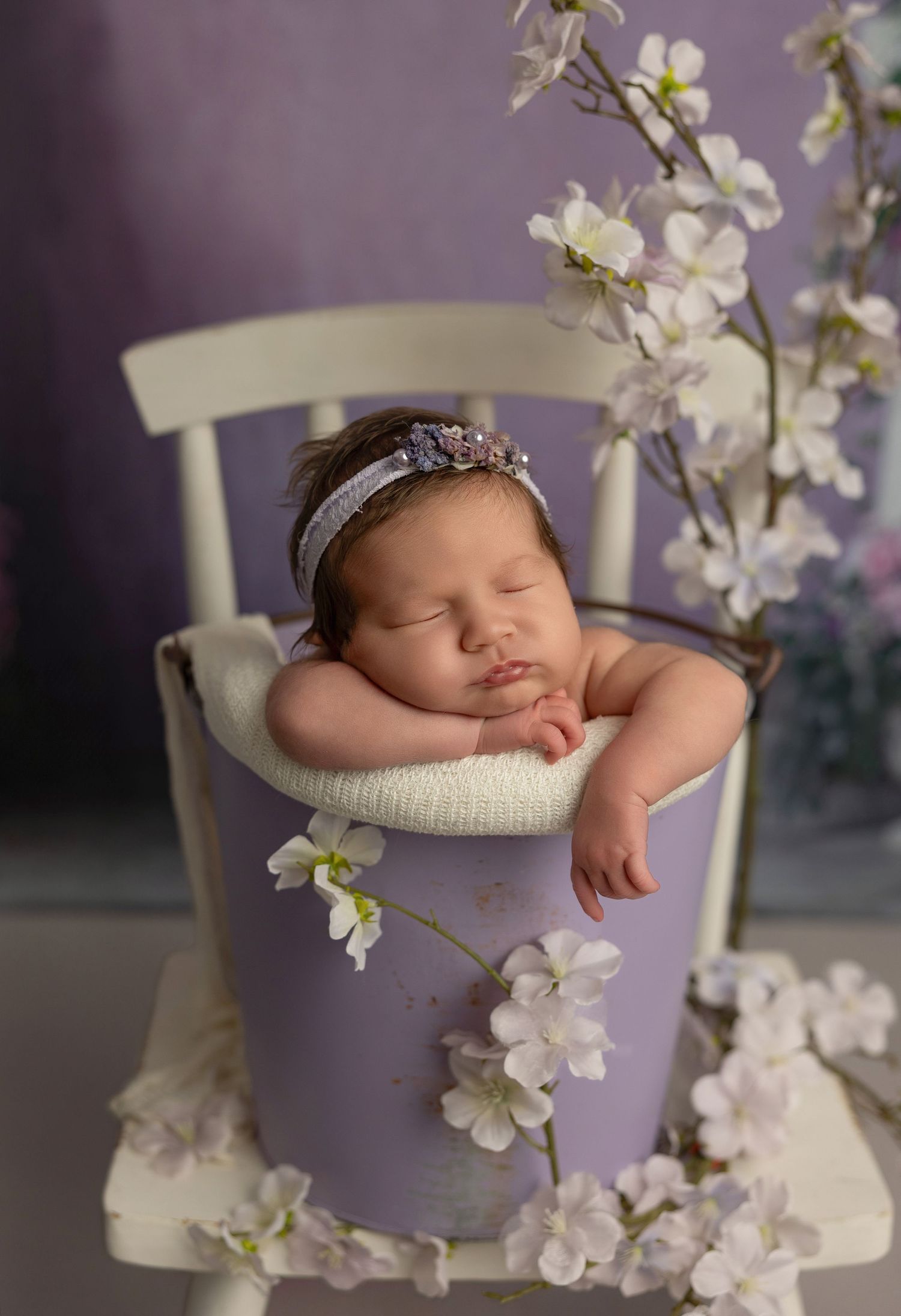 A sleeping newborn nestled in a purple bucket surrounded by delicate white orchid blossoms creates a dreamy spring portrait.