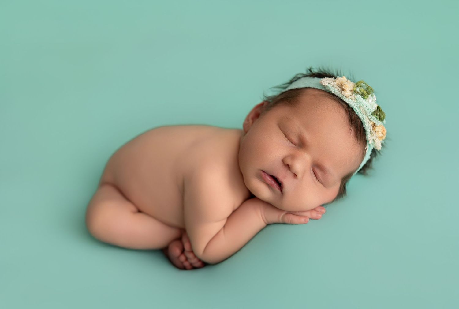 Newborn baby sleeping peacefully on turquoise background wearing delicate floral crown.