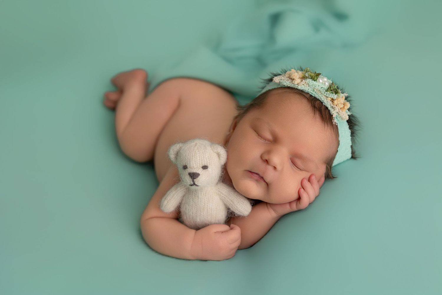 Sleeping newborn wearing floral headband cuddles tiny white teddy bear on mint green backdrop.