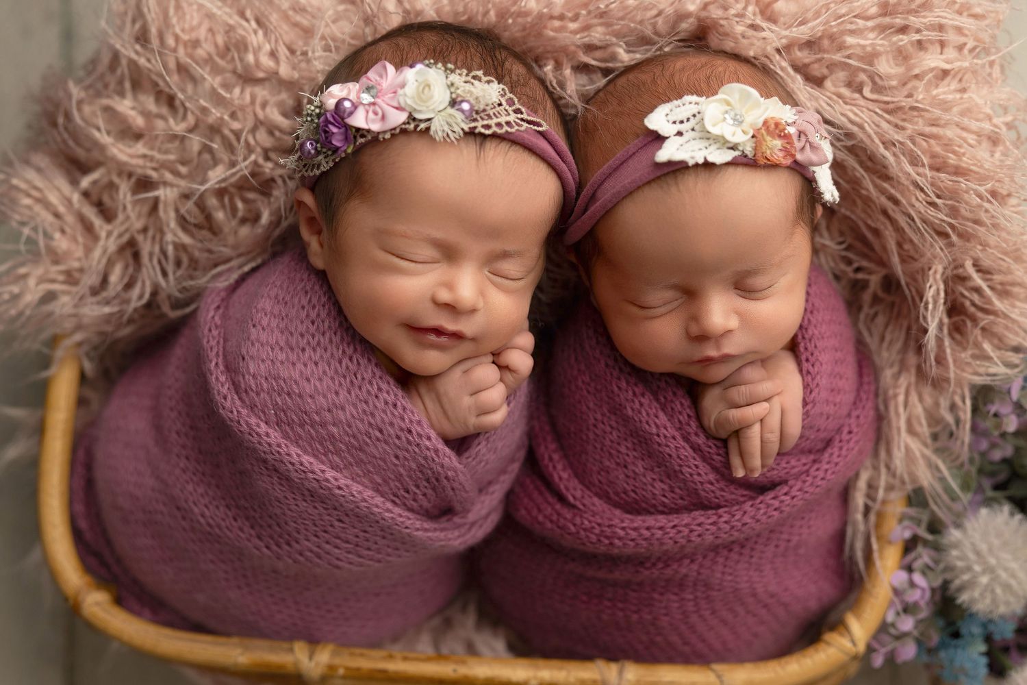 Two sleeping newborns swaddled in purple wraps wearing floral headbands rest together on a fluffy pink backdrop.