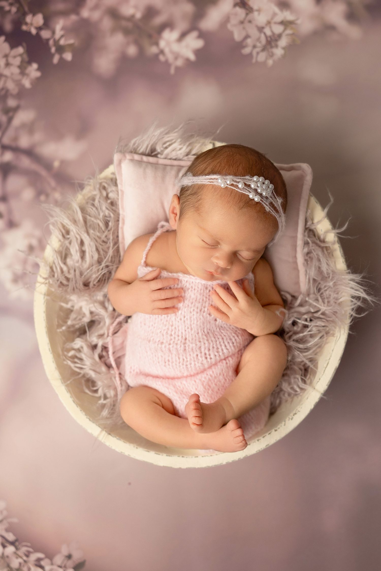 A sleeping newborn wearing pink knit clothing lies peacefully in a white basket surrounded by soft pink cherry blossoms.