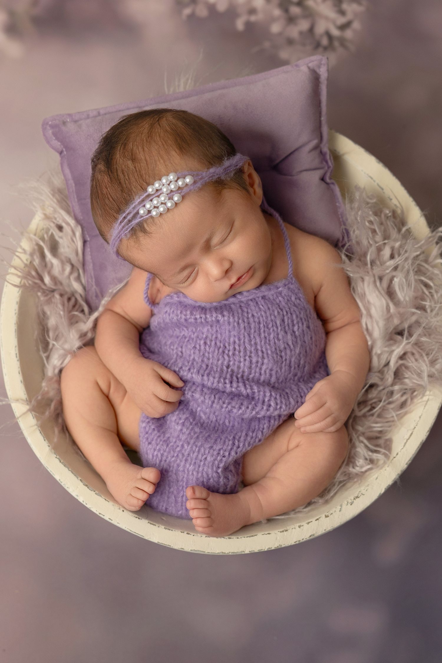 Sleeping newborn wearing purple knit romper and sparkly headband rests peacefully in white bowl on soft gray backdrop.