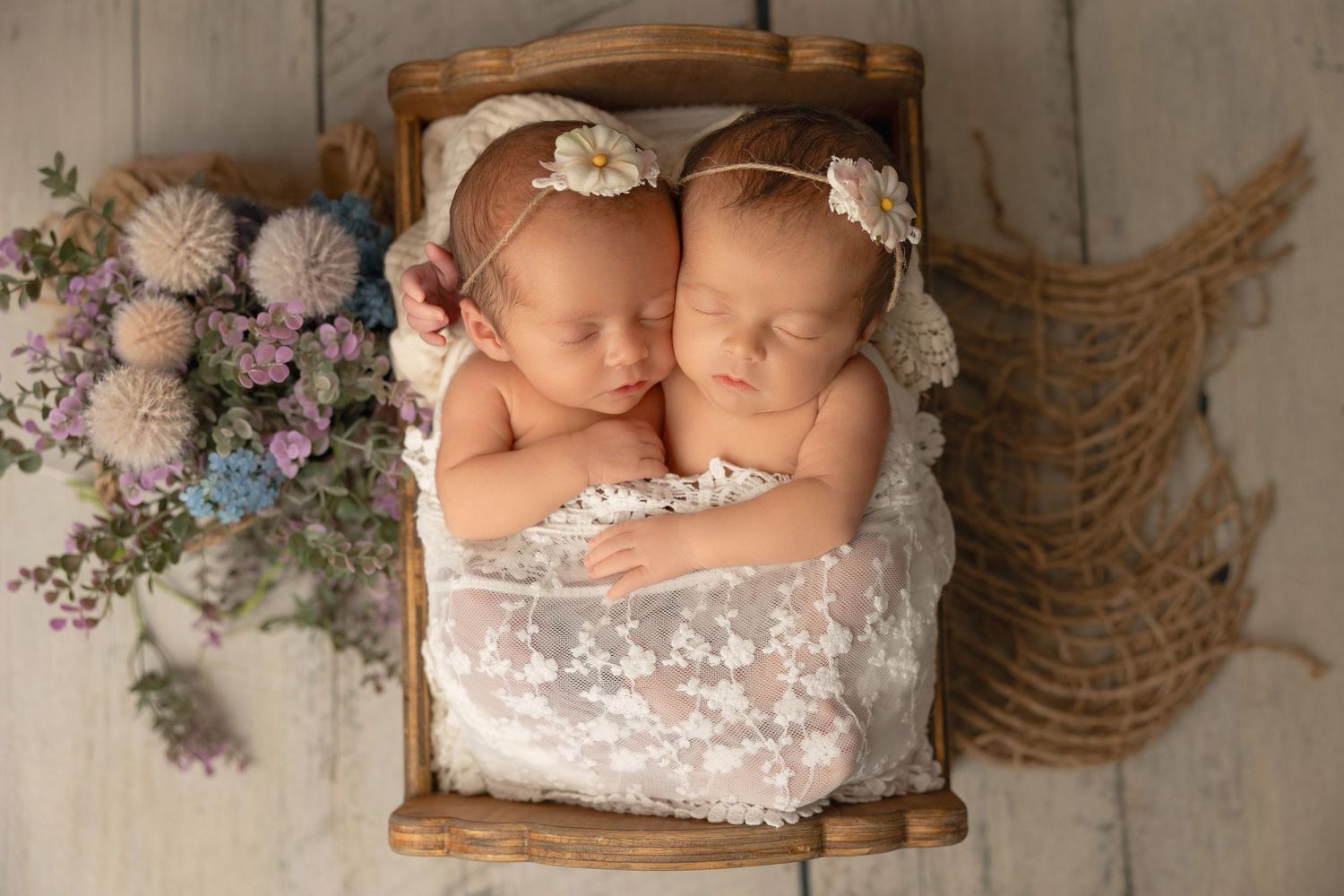 Newborn twins sleeping peacefully in a wooden bed with lace wrap and purple floral decorations.