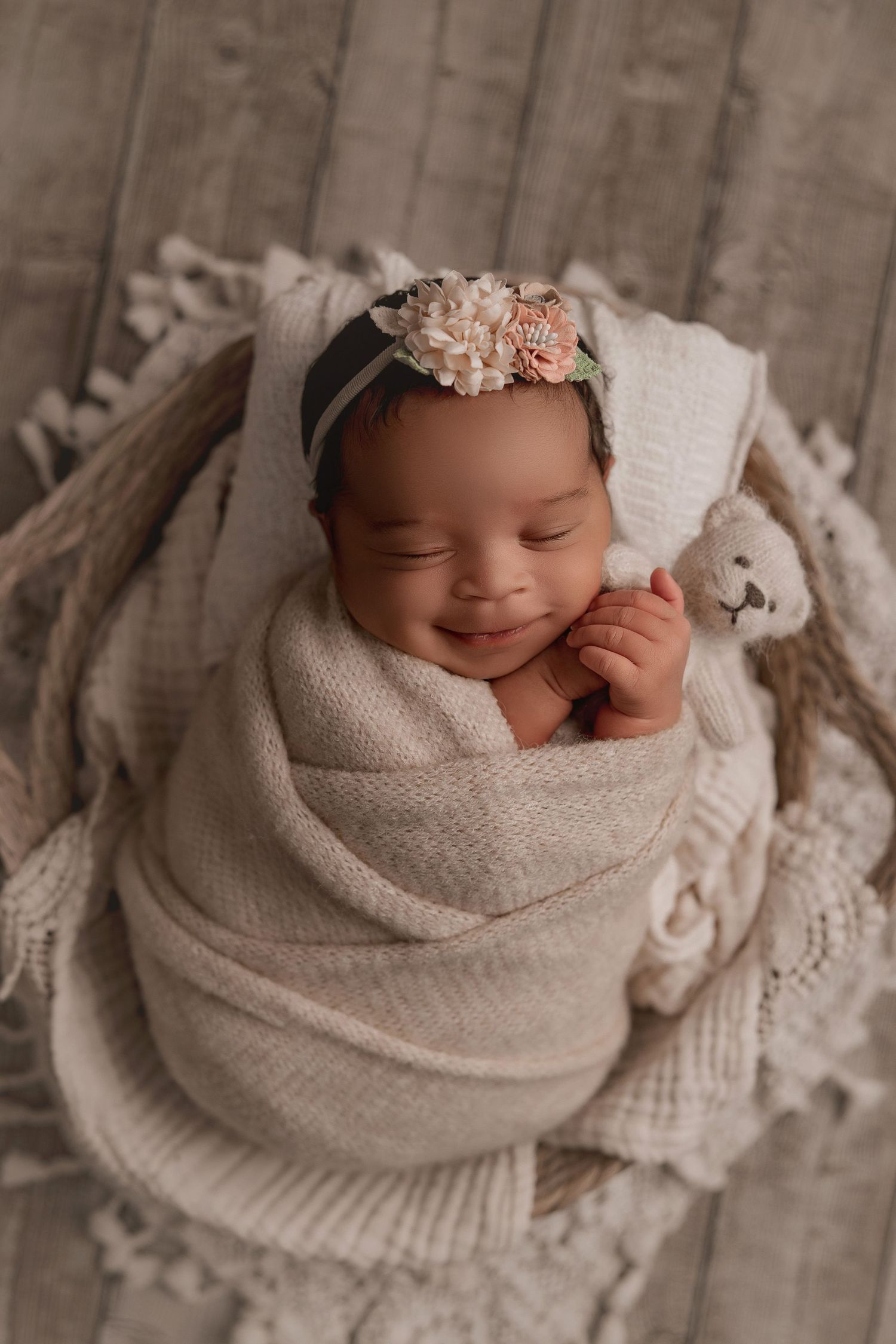 Sleeping newborn wrapped in beige swaddle with floral headband lying on textured blanket in studio setting.