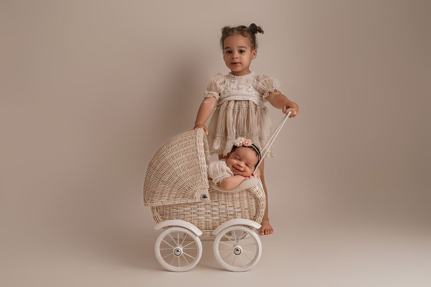 Two children in vintage-style lace dresses pose with an antique white wicker baby carriage against a neutral backdrop.