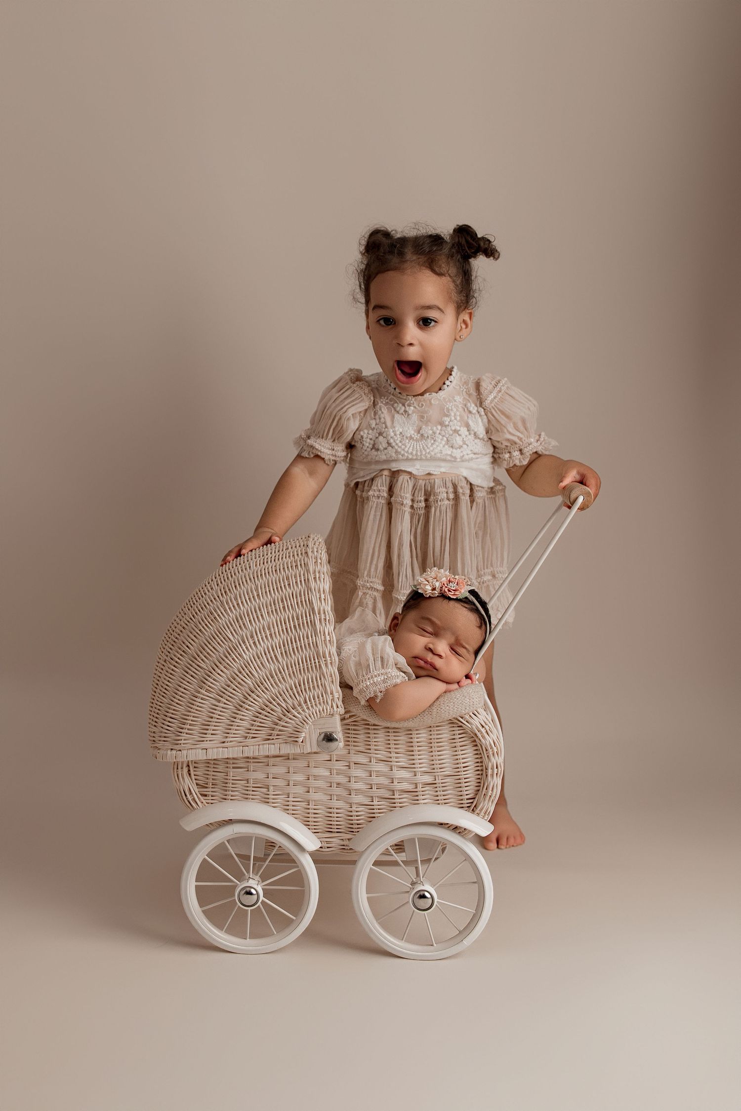 A vintage-style portrait featuring a white wicker doll carriage with two children in light-colored clothing against a neutral backdrop.