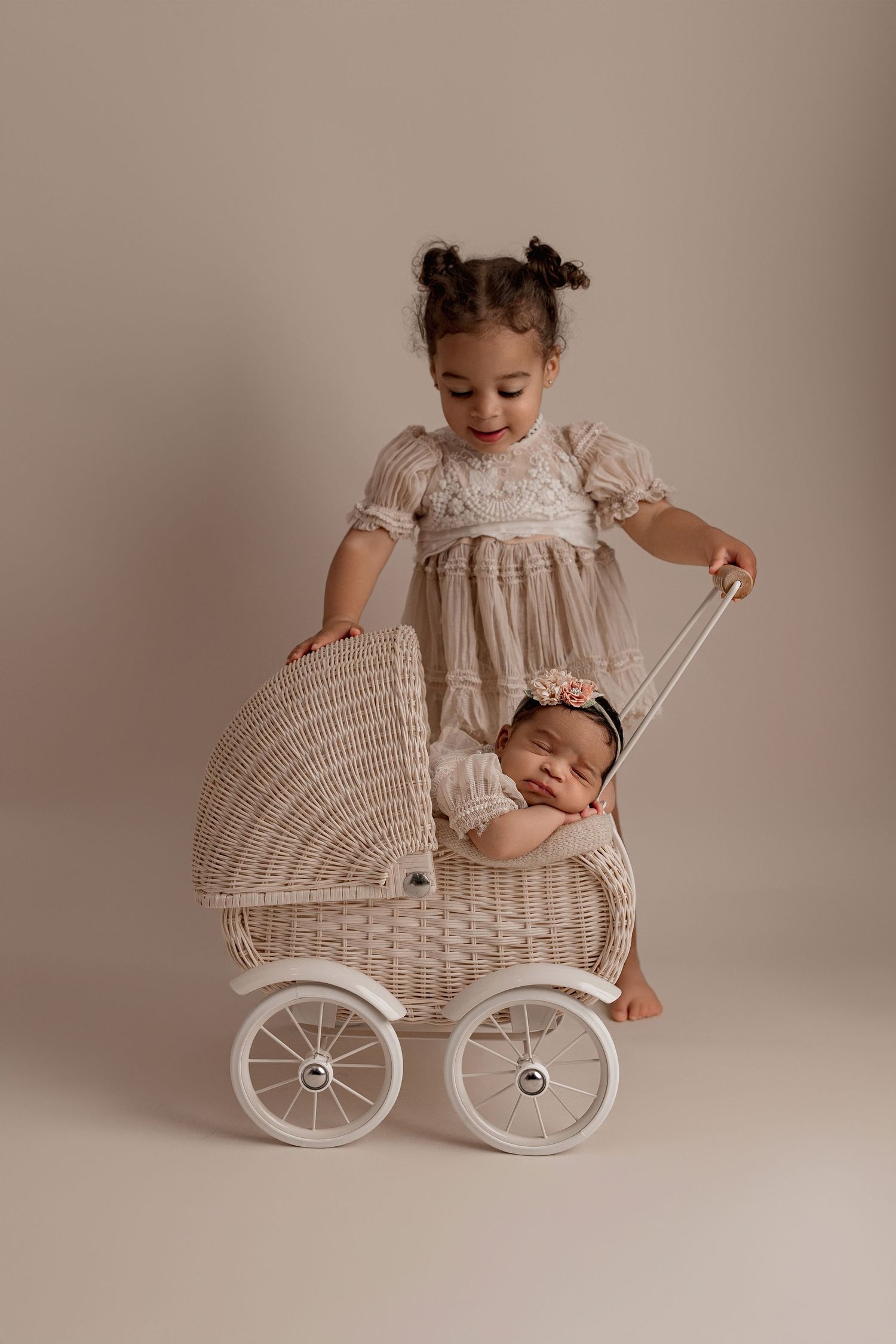 Two children with a vintage white wicker baby carriage in a soft neutral studio setting.