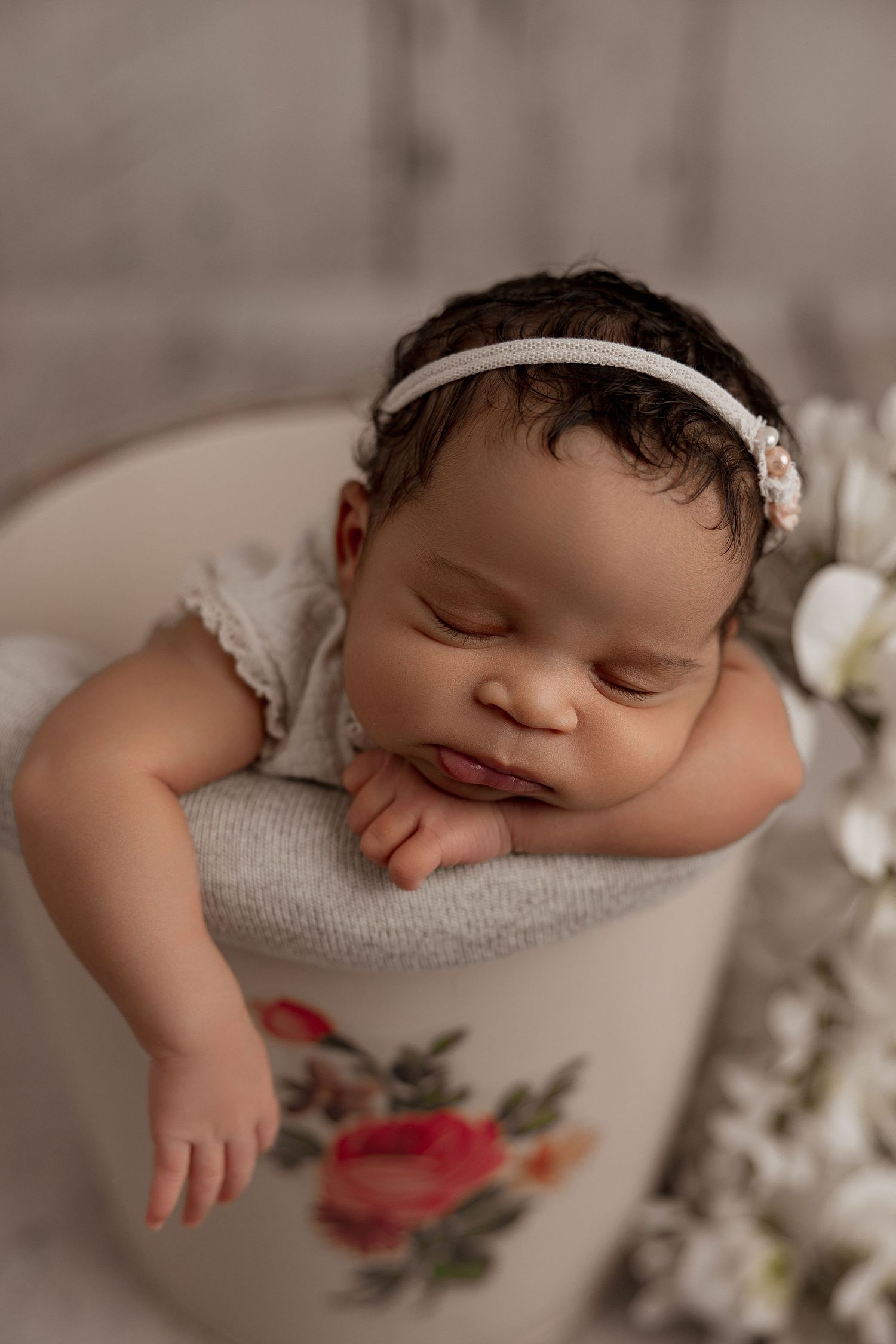 Sleeping baby with headband resting in a decorative floral container during newborn photoshoot.