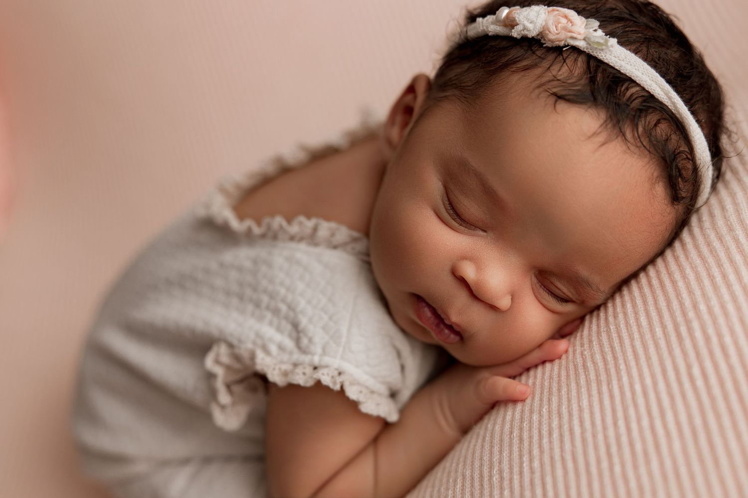 Peaceful newborn wearing white dress and delicate headband resting on soft pink backdrop.