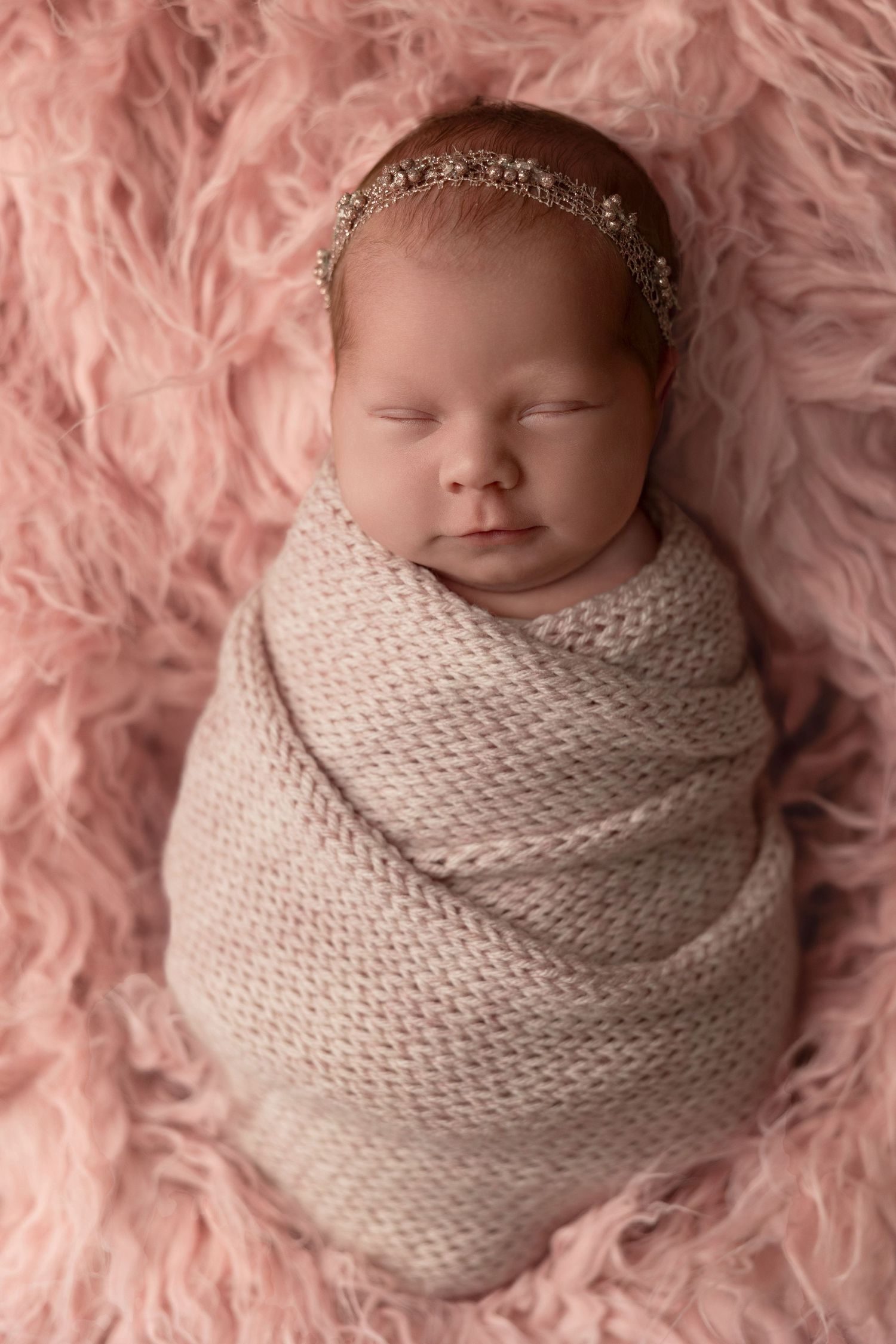 Sleeping newborn wrapped in soft white knit blanket wearing delicate headband on pink furry backdrop.