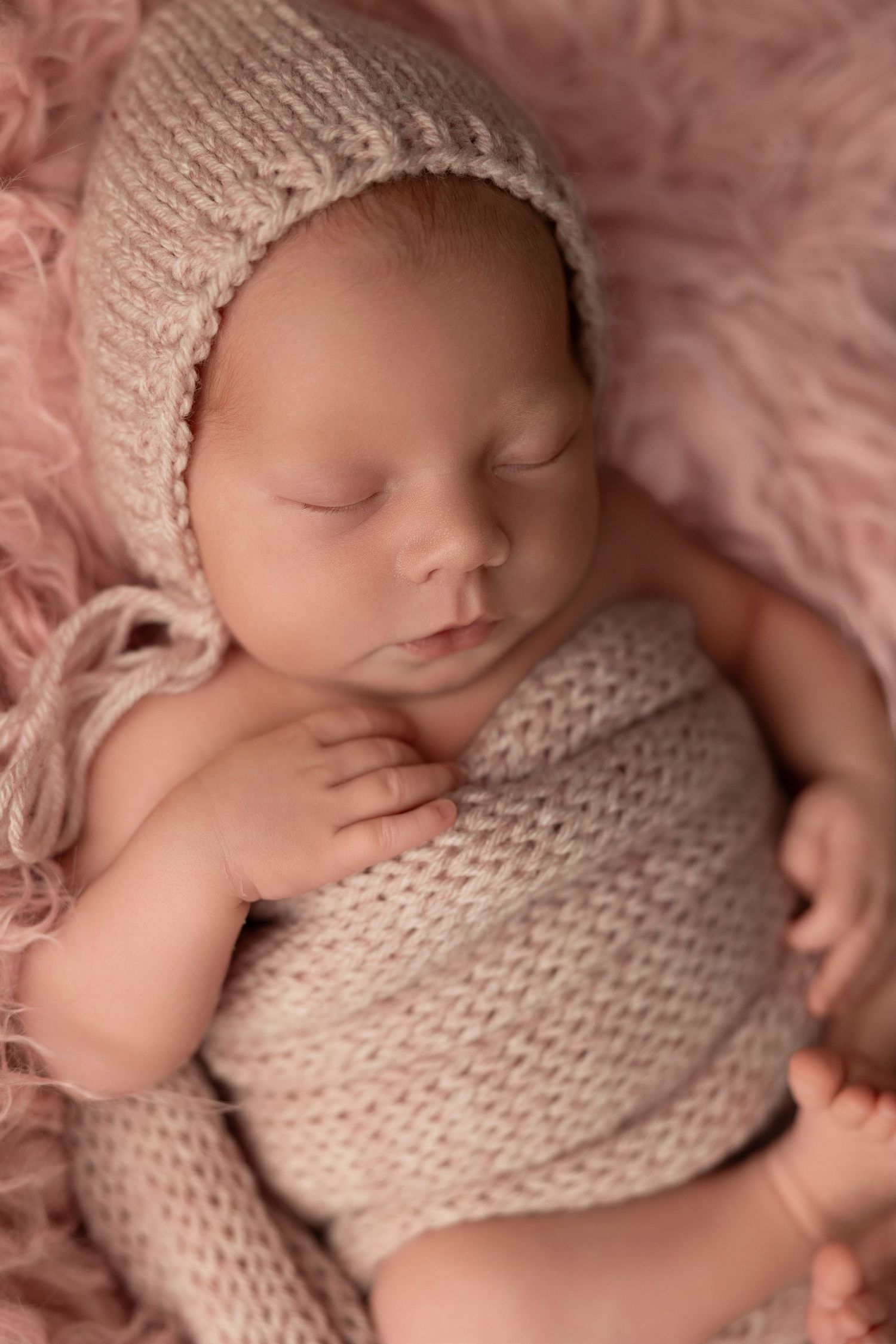 Sleeping newborn wearing soft knitted beige bonnet and romper on pink blanket in studio portrait setting.