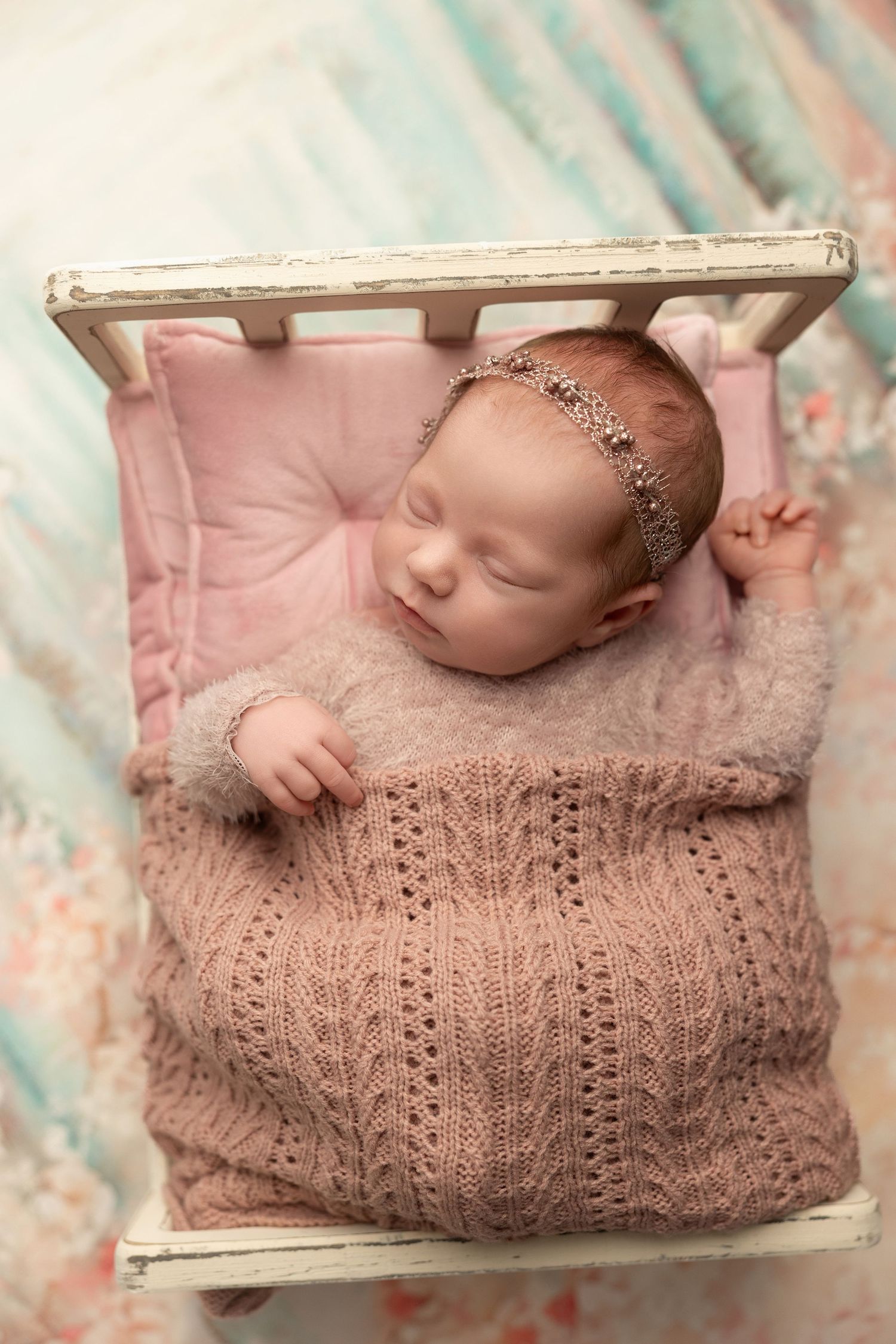 Newborn baby sleeping peacefully in vintage white drawer on pink tufted cushion wearing delicate headband and knit blanket.