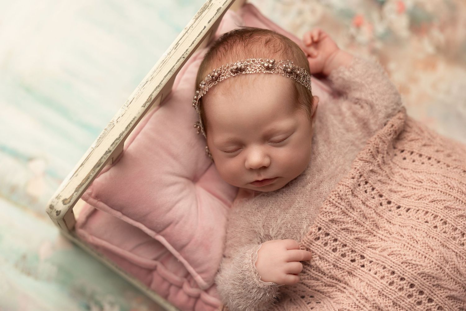 Sleeping newborn wrapped in soft pink knitted blanket wearing delicate headband on pastel background.