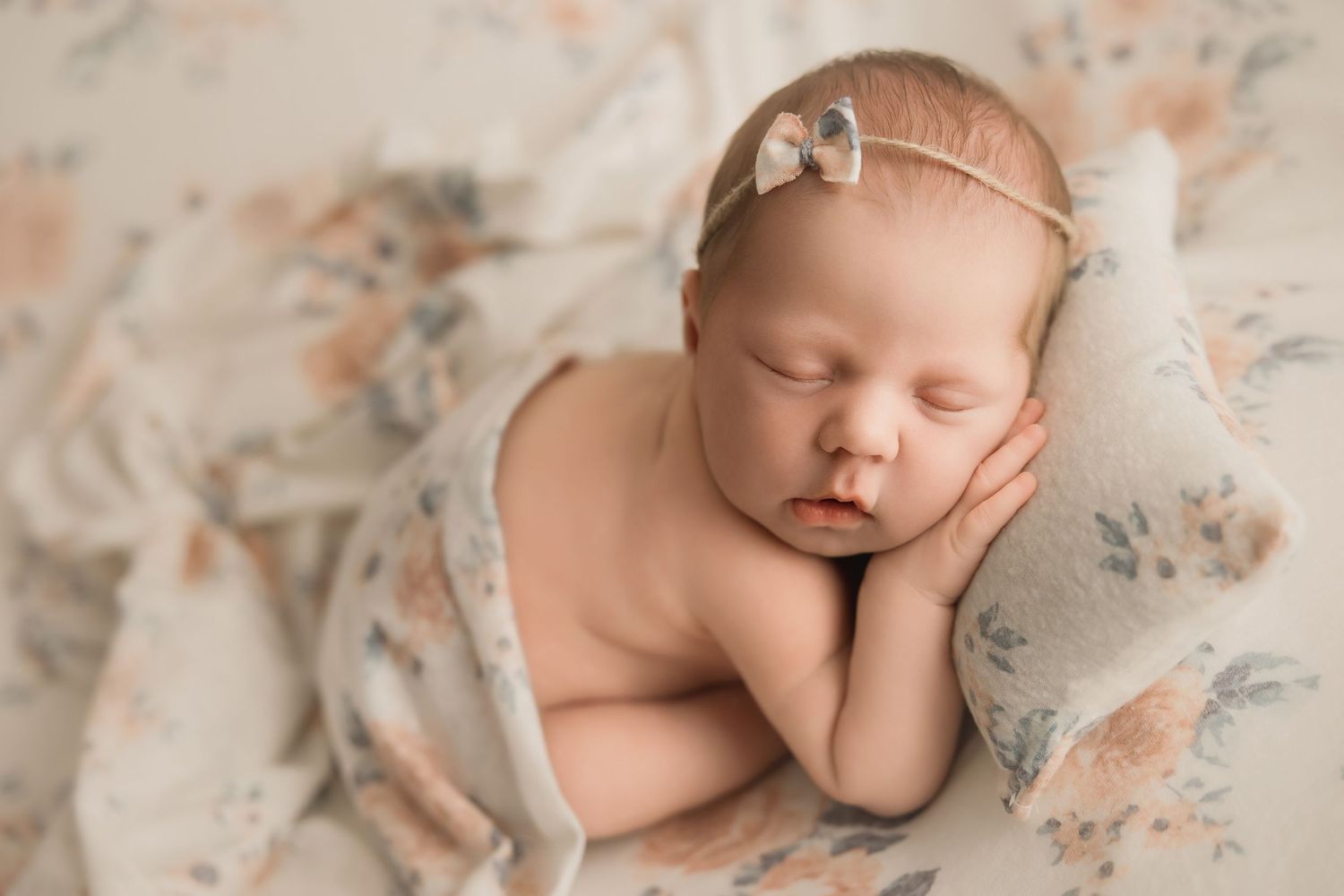 A peaceful sleeping newborn rests on soft floral fabric wearing a delicate bow headband.
