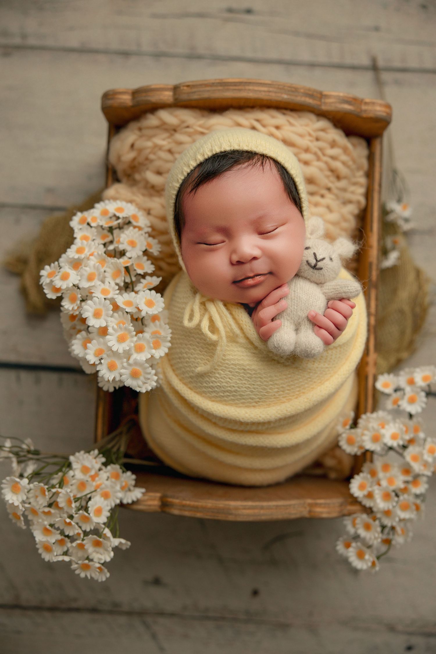 Smiling newborn baby swaddled in yellow knit wrap, sleeping in wooden crib with daisy flowers and teddy bear.