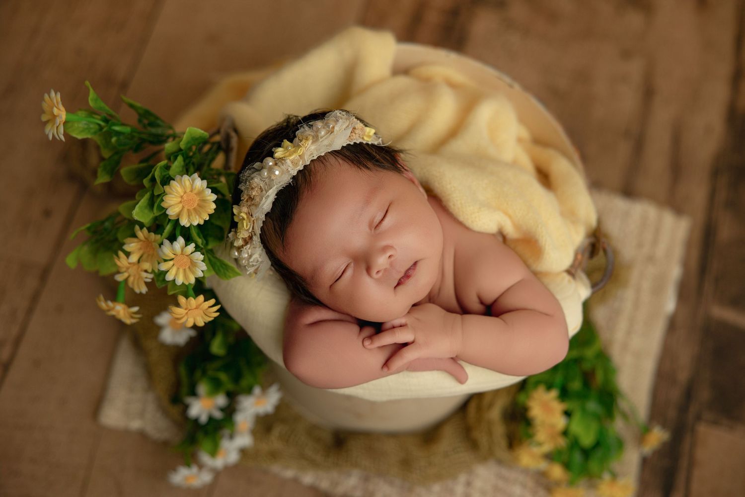 Newborn baby sleeping peacefully in a wooden bowl with yellow daisy flowers and soft yellow blanket.