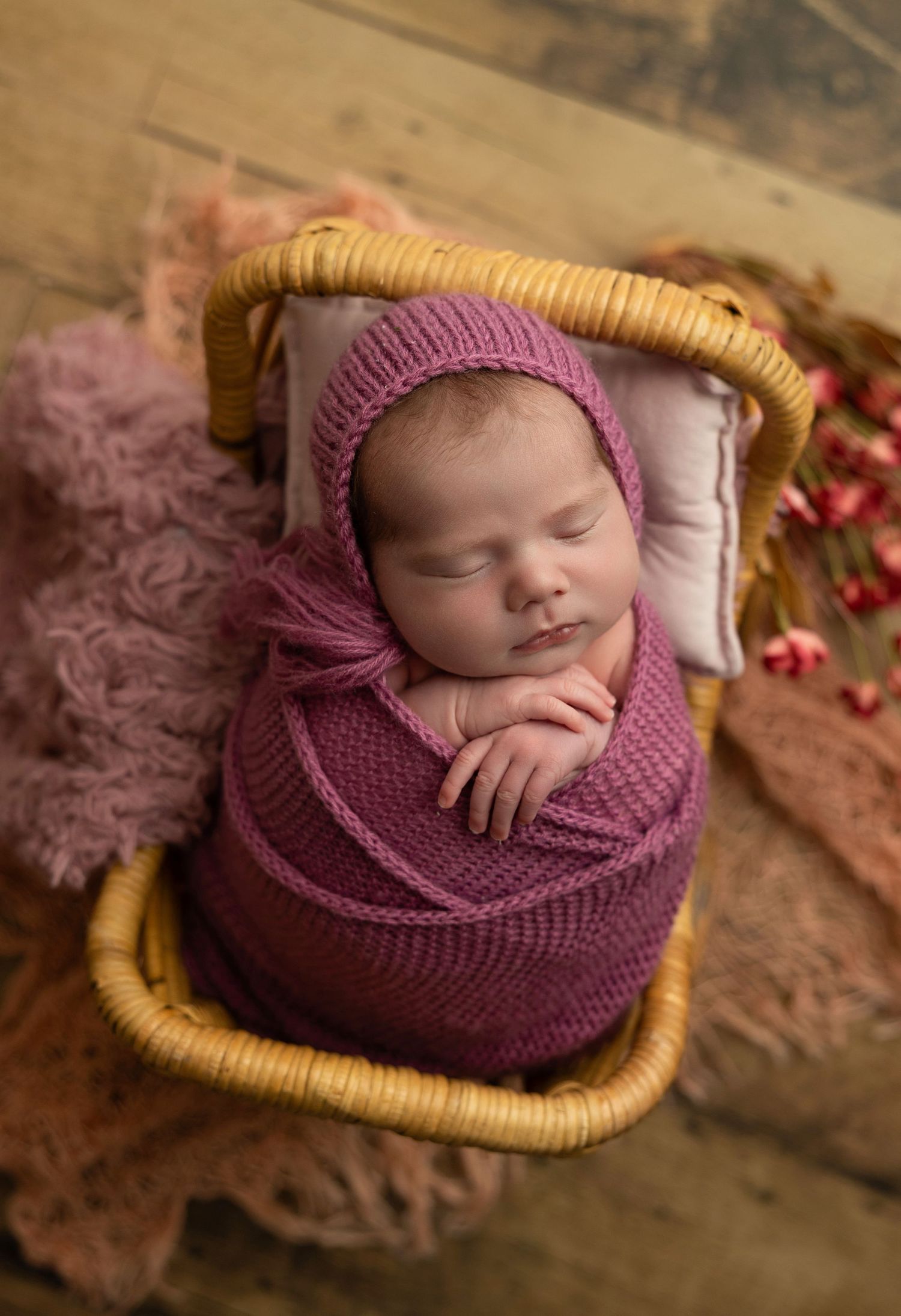 Sleeping newborn wrapped in purple knit blanket nestled in woven basket with rustic wood background.