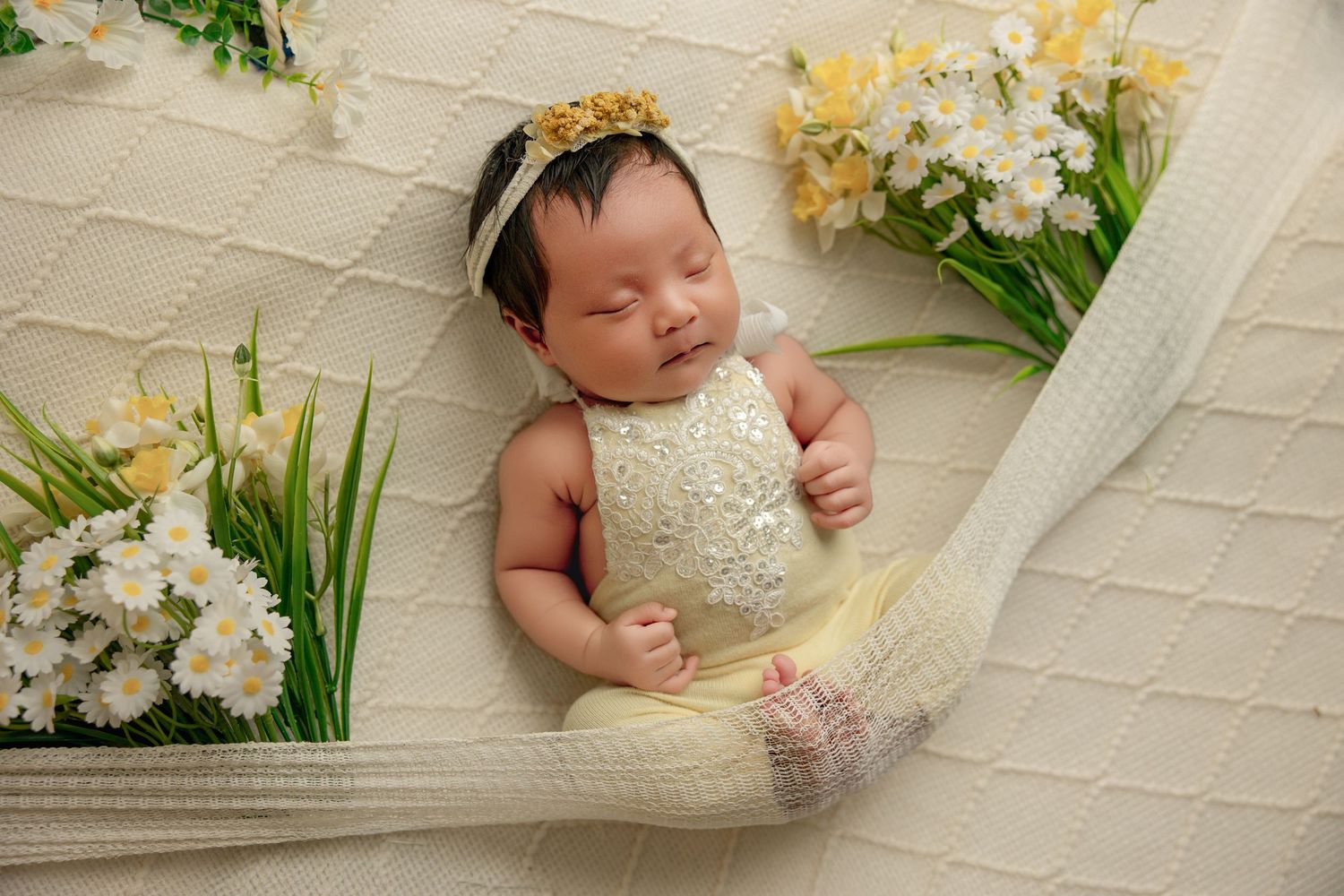Newborn baby sleeping peacefully in a white knit hammock surrounded by yellow and white flowers wearing a floral headband.