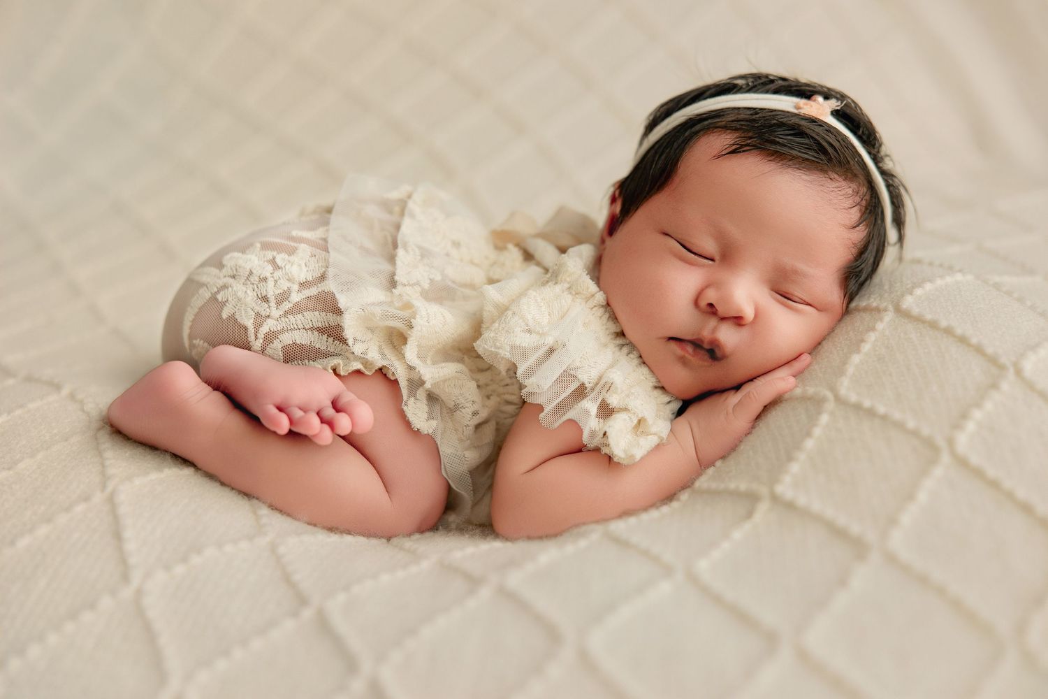 Newborn baby girl sleeping peacefully in a white lace romper on a cream textured blanket with a floral headband.
