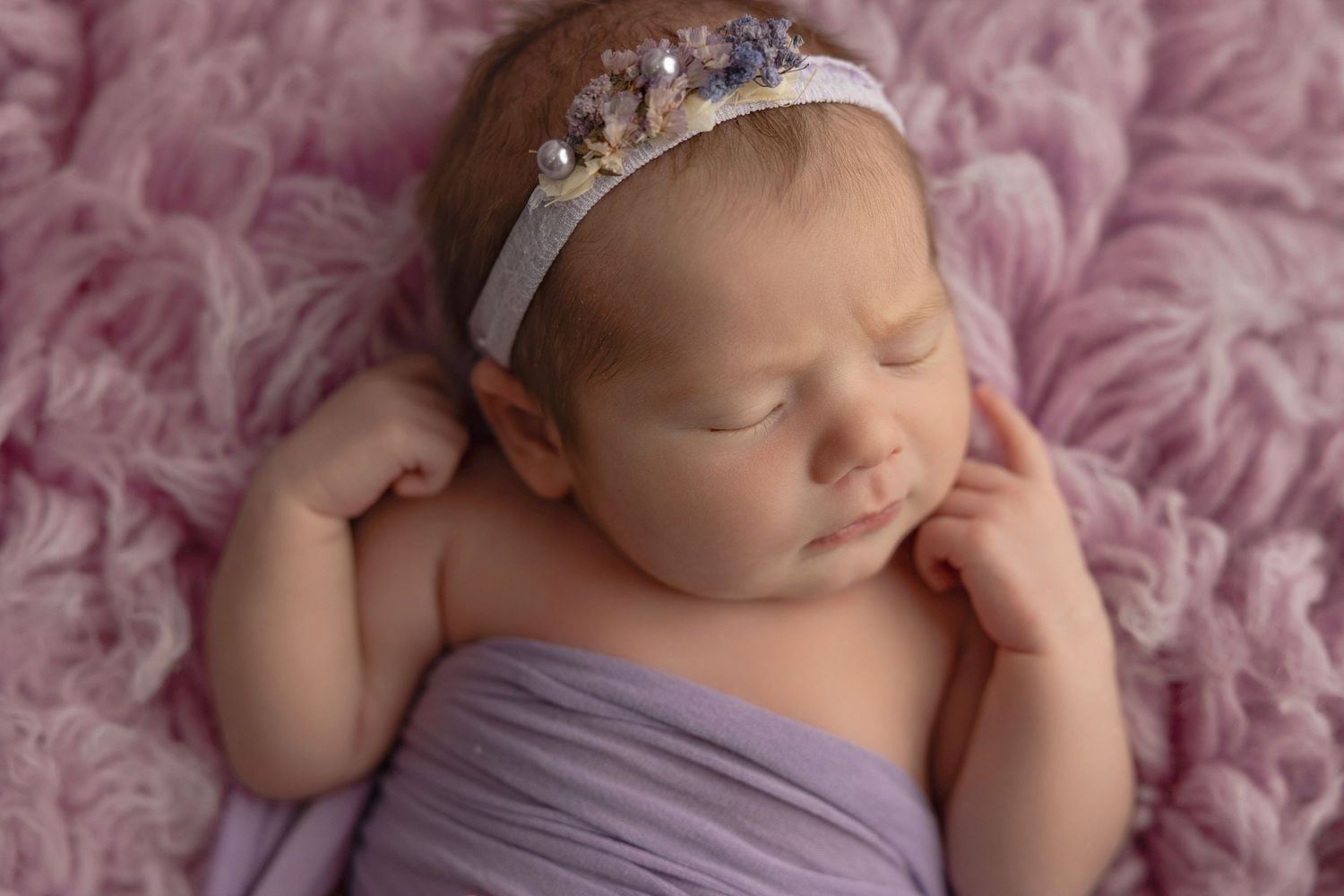 Sleeping newborn wrapped in purple fabric wearing delicate crown headband on pink textured blanket.