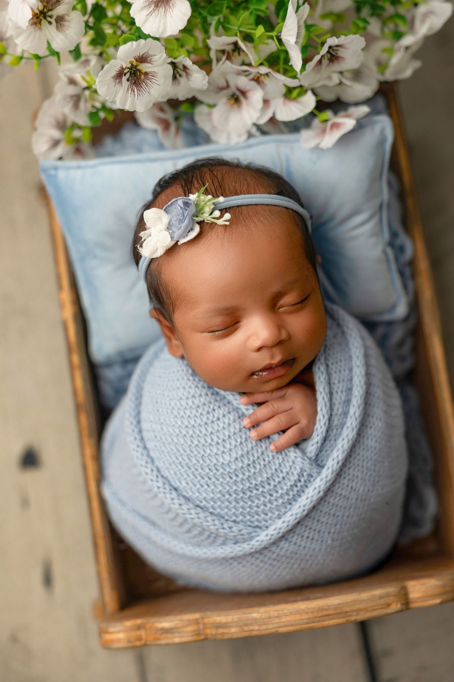 Sleeping newborn baby girl swaddled in blue knit wrap with floral headband in wooden crate with flowers.