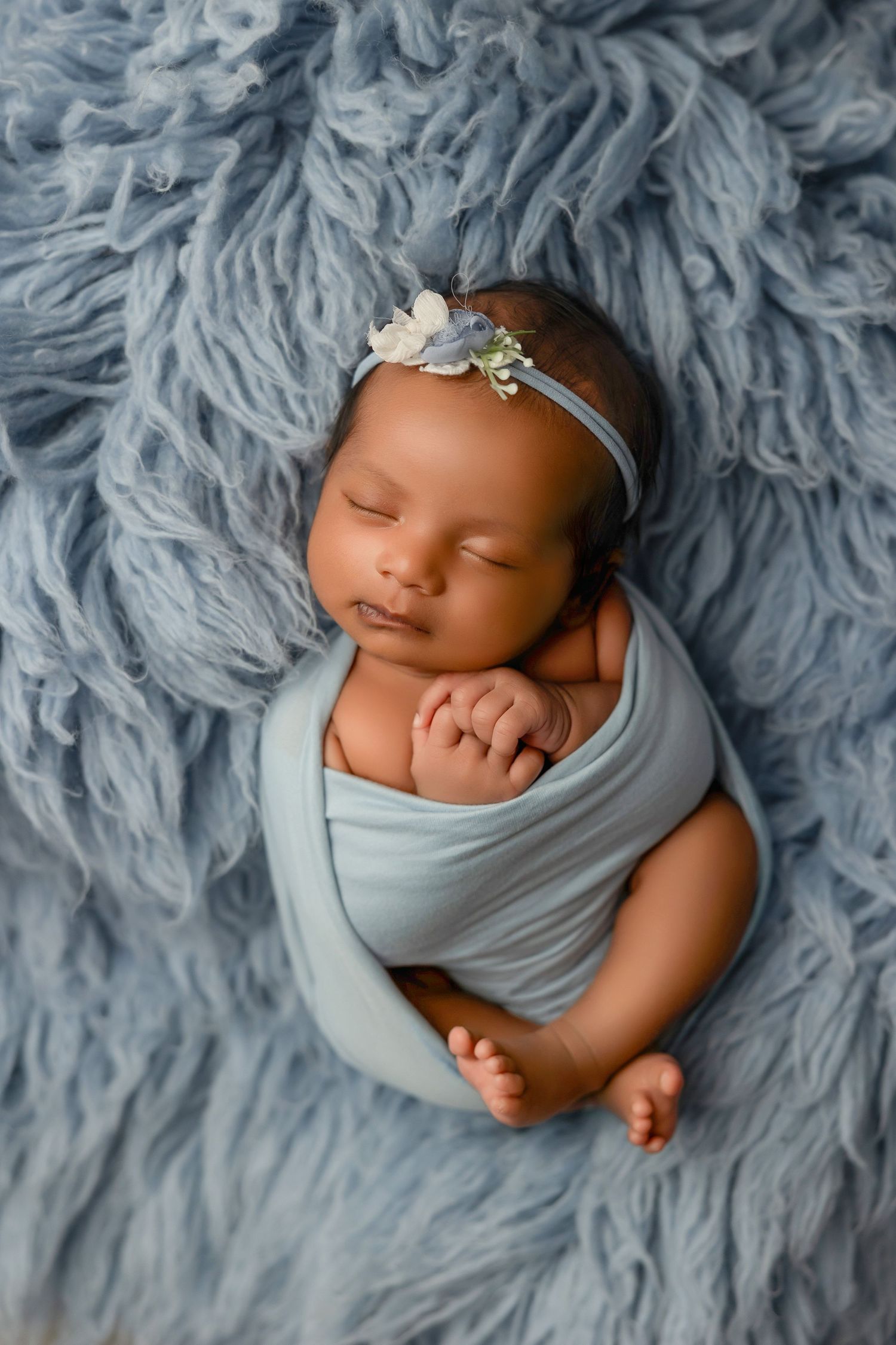 Newborn baby girl swaddled in blue wrap with floral headband sleeping peacefully on a soft blue fluffy blanket.