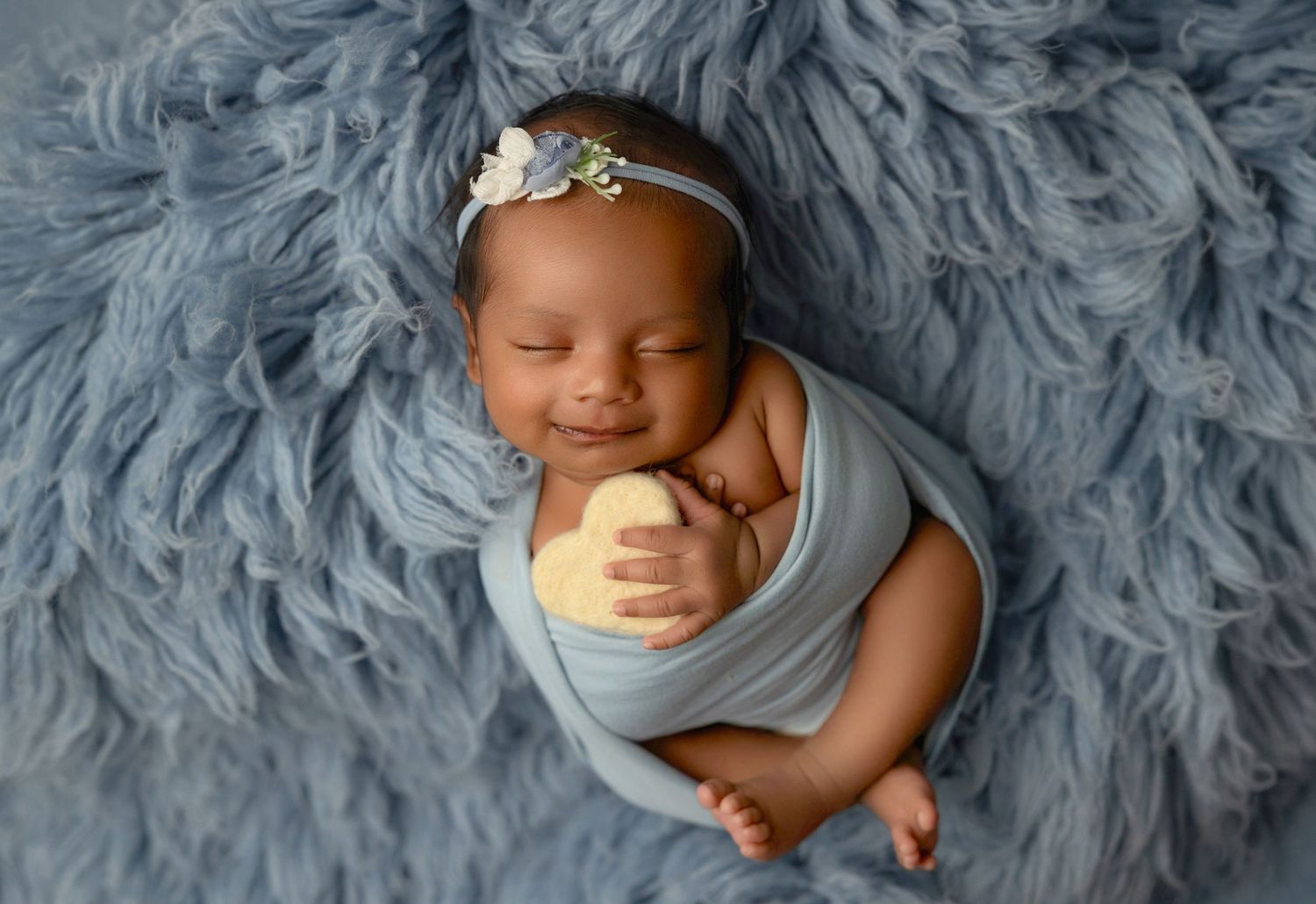 Smiling newborn baby wrapped in blue swaddle with floral headband resting on soft blue fluffy blanket.