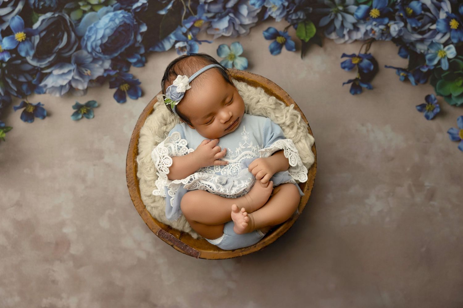 Newborn baby girl sleeping in wooden bowl wearing lace outfit with floral headband surrounded by blue flowers.