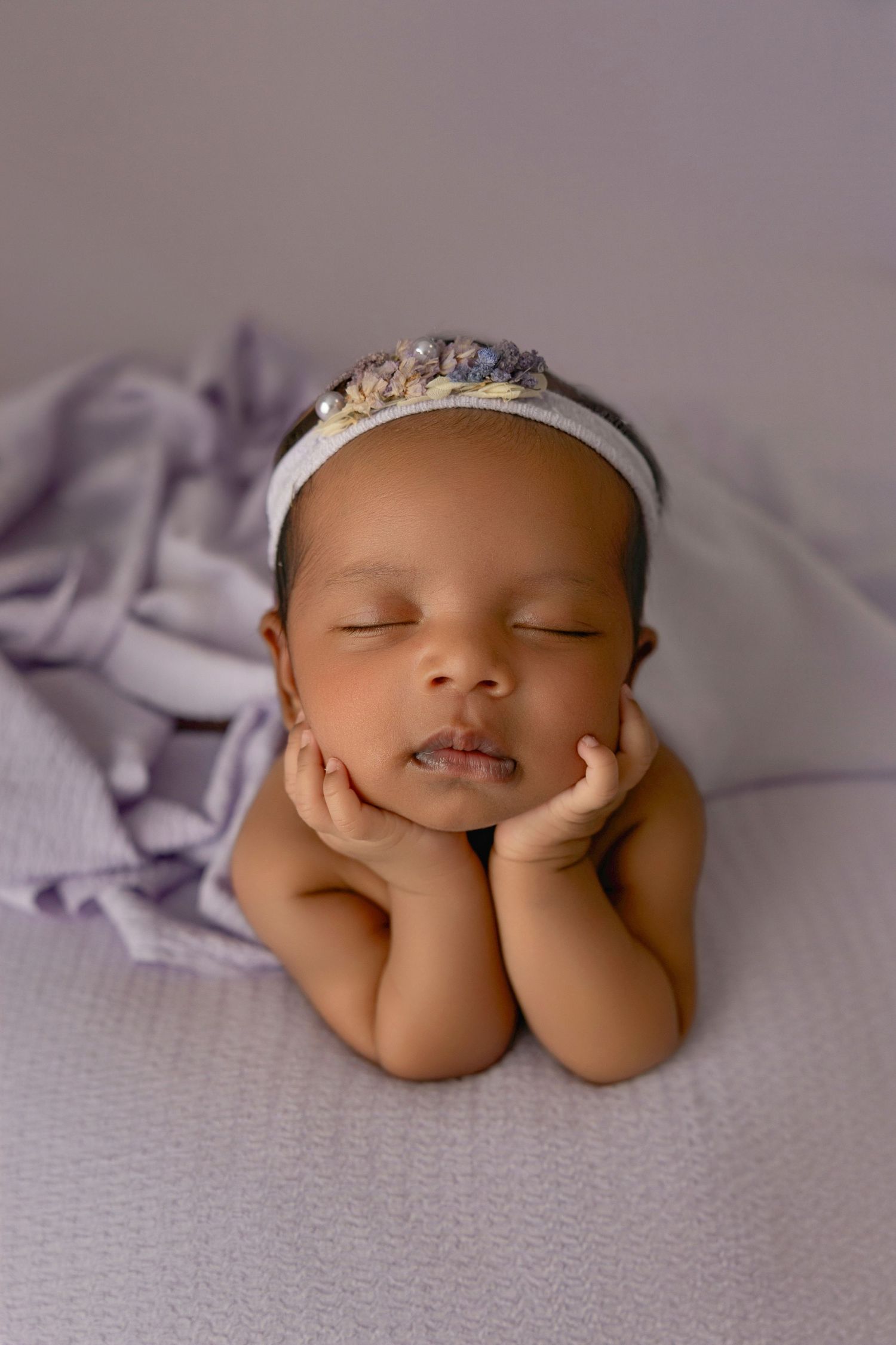 Newborn baby girl sleeping peacefully with hands on cheeks, wearing a delicate floral headband on a soft lavender background.