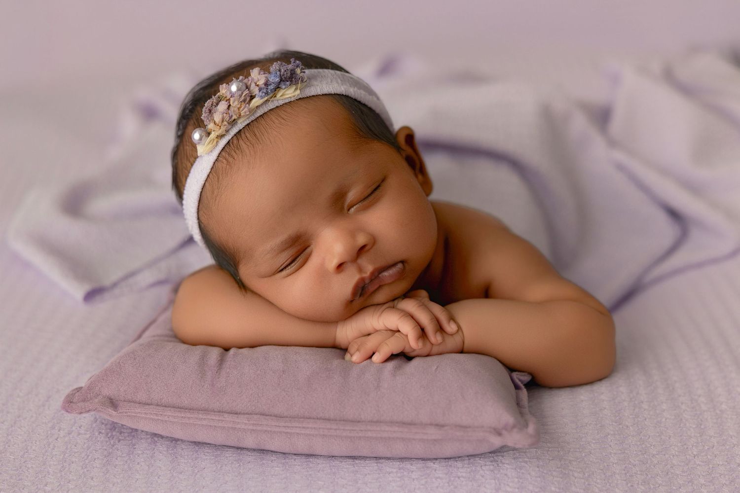 Sleeping newborn baby girl wearing a floral headband resting on a lavender pillow in a purple-themed photo.