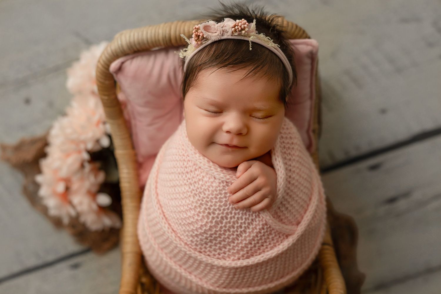 Newborn baby wrapped in soft pink knit blanket sleeping peacefully in wooden basket on rustic floor.