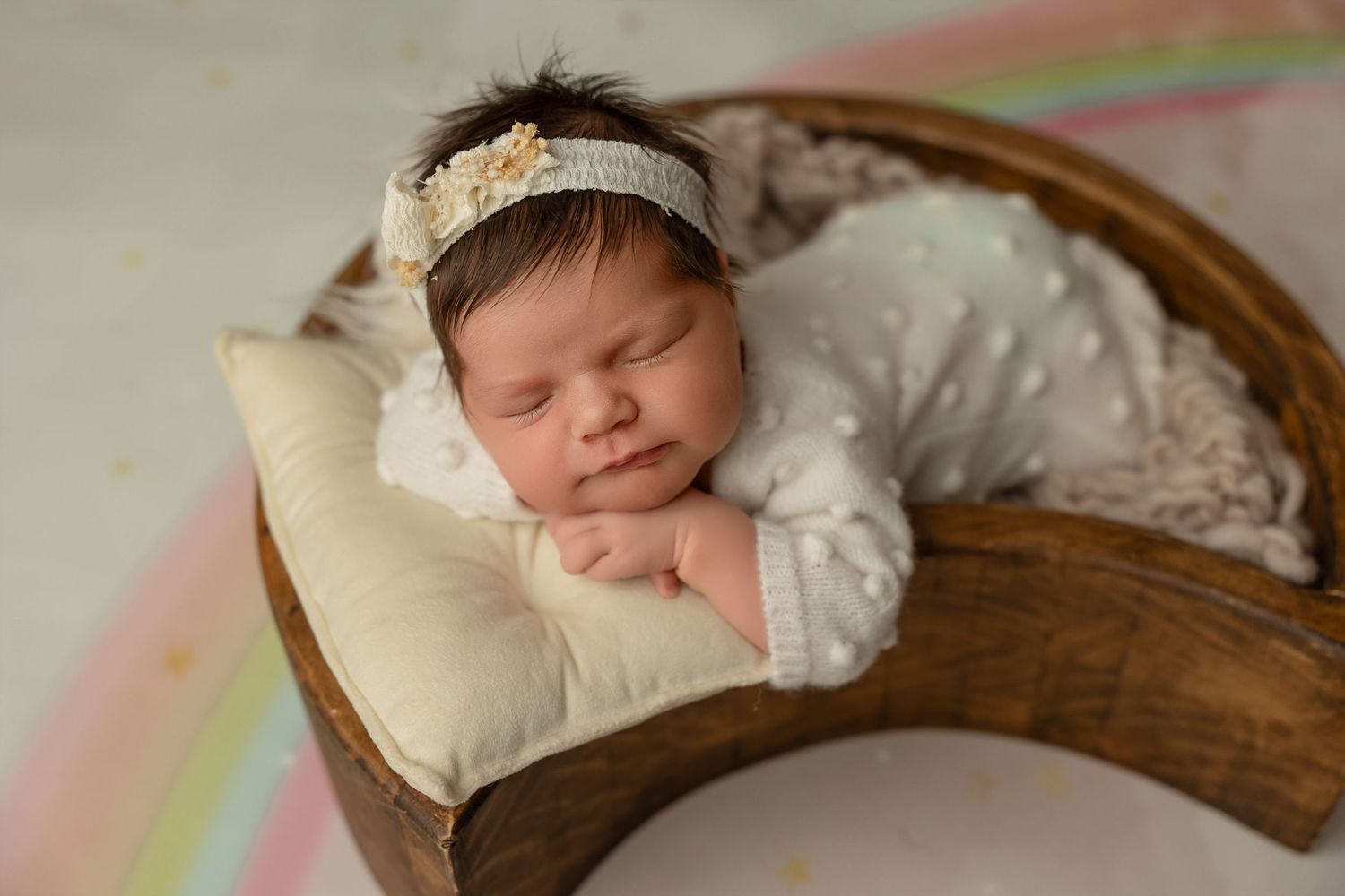 Sleeping newborn in white lace dress rests peacefully on cream pillow in rustic wooden basket against pastel backdrop.