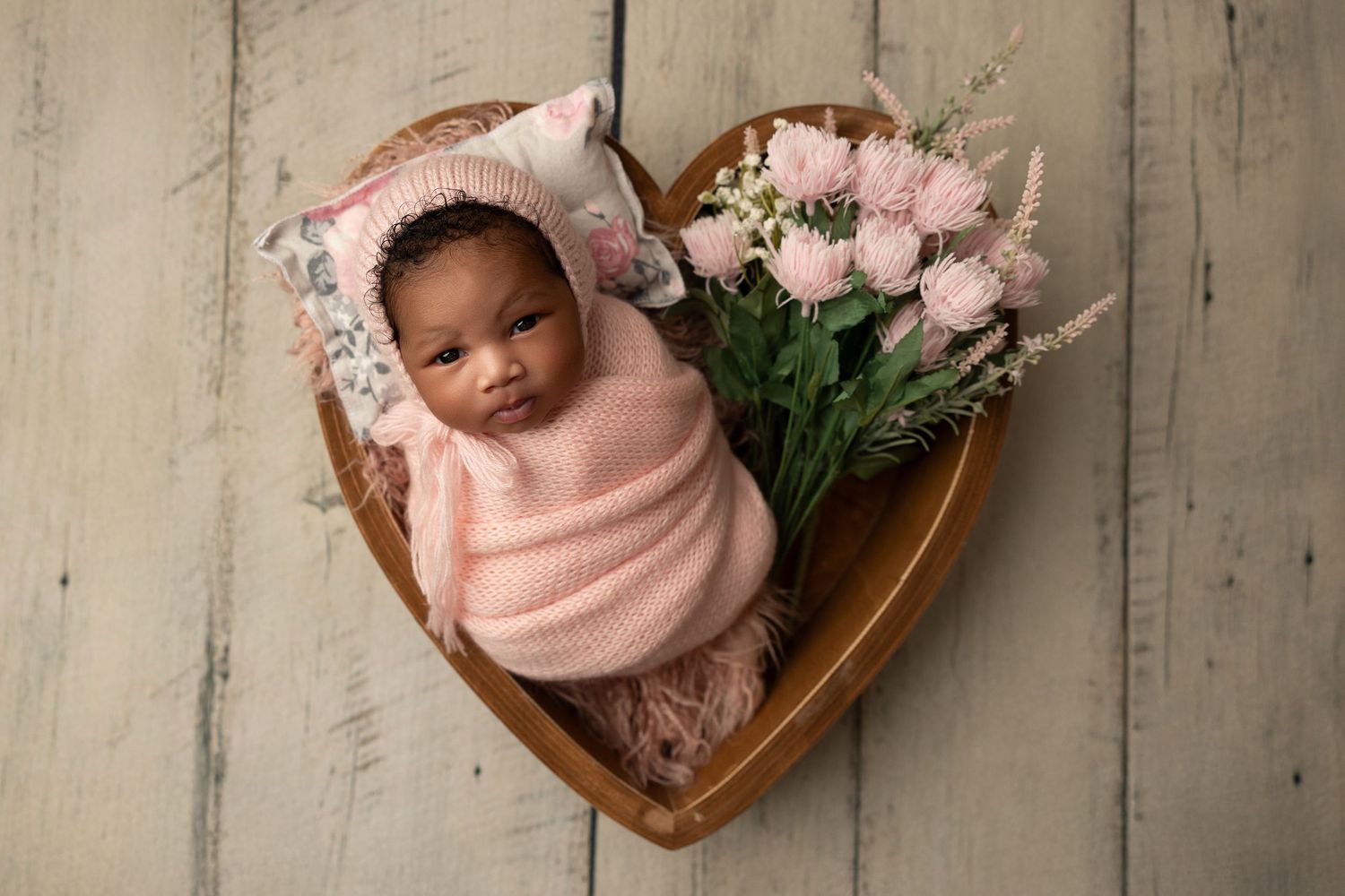 Newborn resting in heart-shaped wooden bowl with pink flowers.