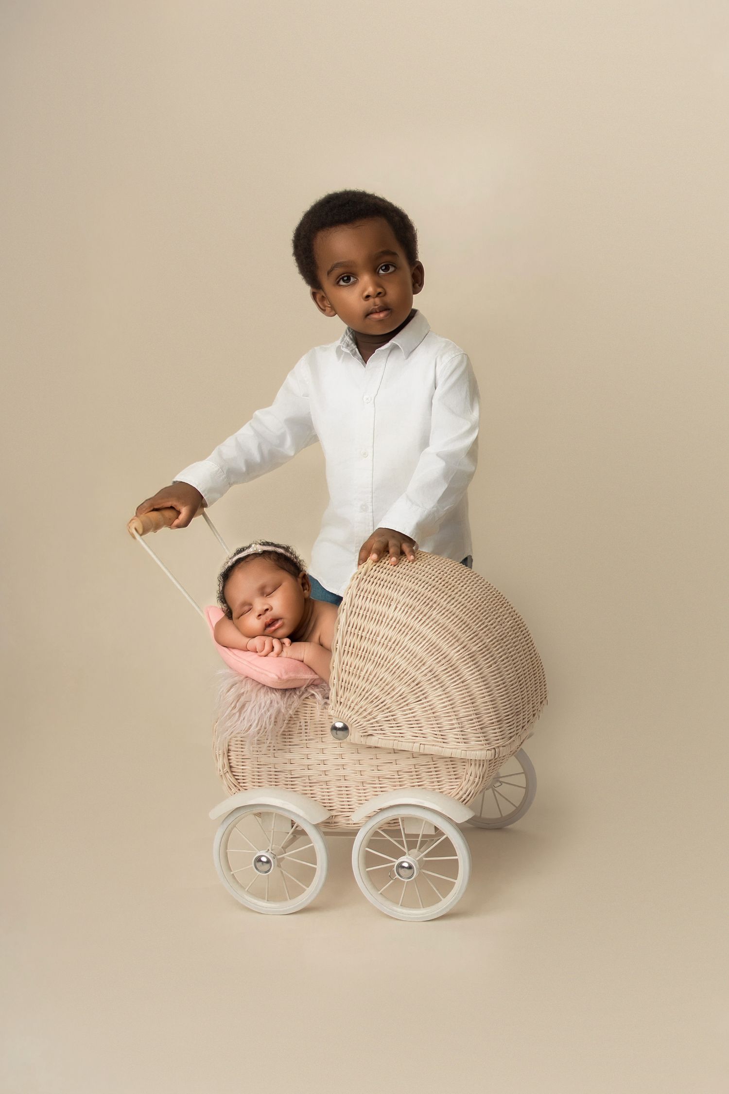 A baby in a white outfit sits in a vintage-style wicker carriage against a neutral beige background.