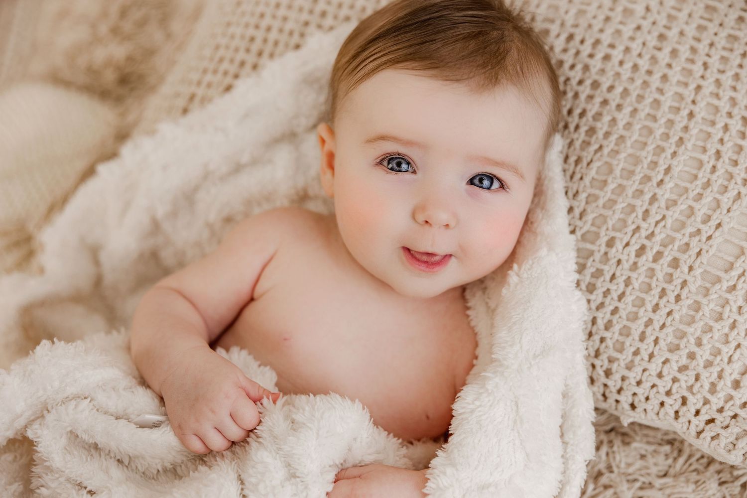 A baby wrapped in soft beige fabric lies on textured burlap material during a newborn photography session.