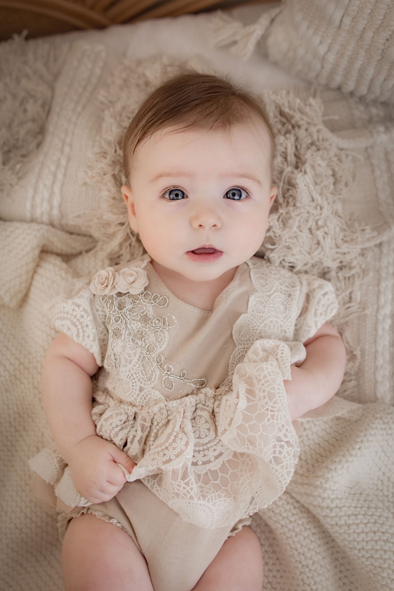 Baby wearing delicate cream lace dress lies on soft knit blanket in gentle natural lighting.