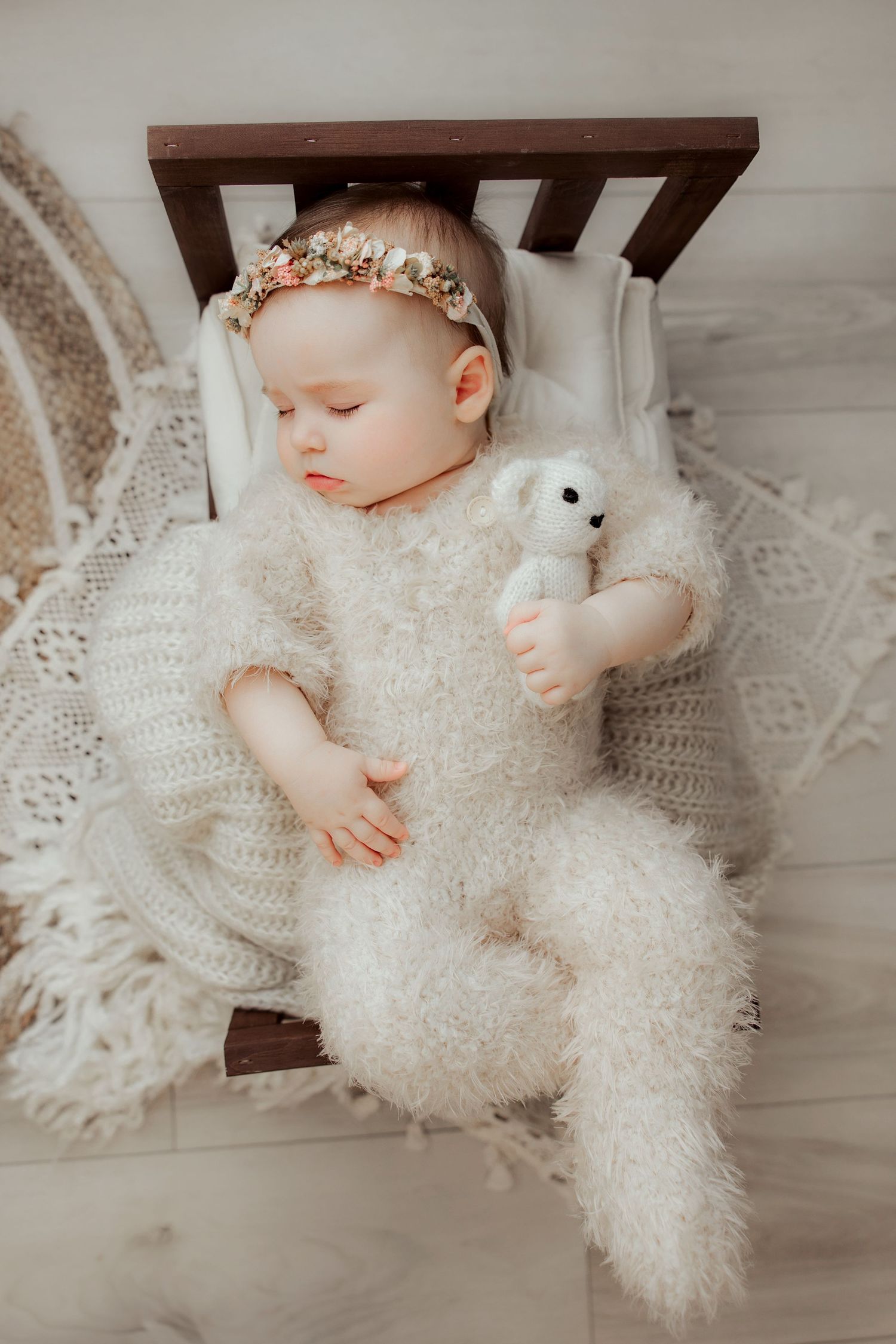 A sleeping baby in a delicate white lace dress lies on a light wooden floor wearing a small crown accessory.