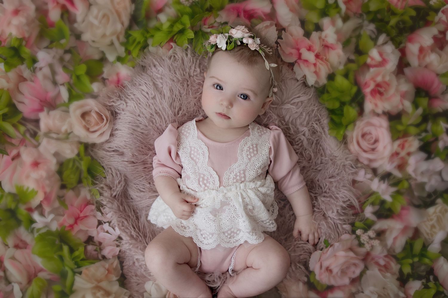 A baby wearing a white lace dress and floral crown lies on a furry blanket surrounded by pink roses and greenery.