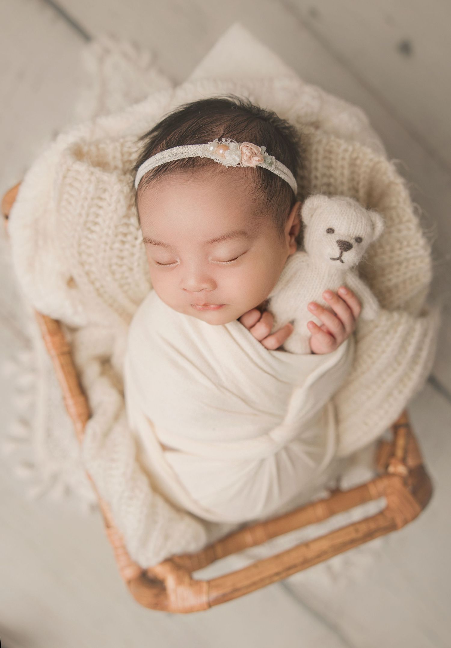 A sleeping newborn wrapped in white knit blankets rests peacefully in a rustic wooden basket.