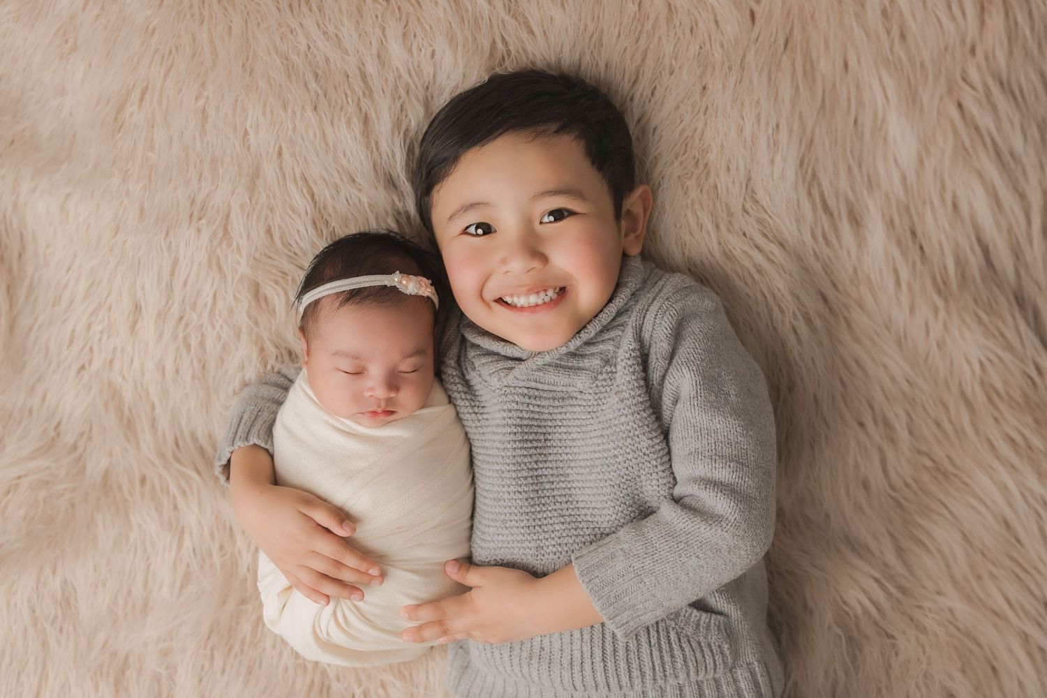 Two children in gray sweaters lying on beige carpet, with one cradling a swaddled baby.