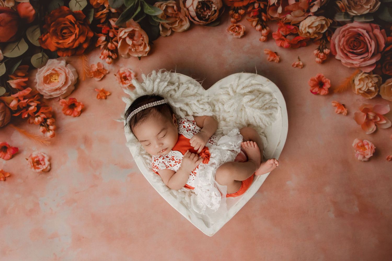 Newborn baby sleeping in heart-shaped basket surrounded by coral roses on pink background.