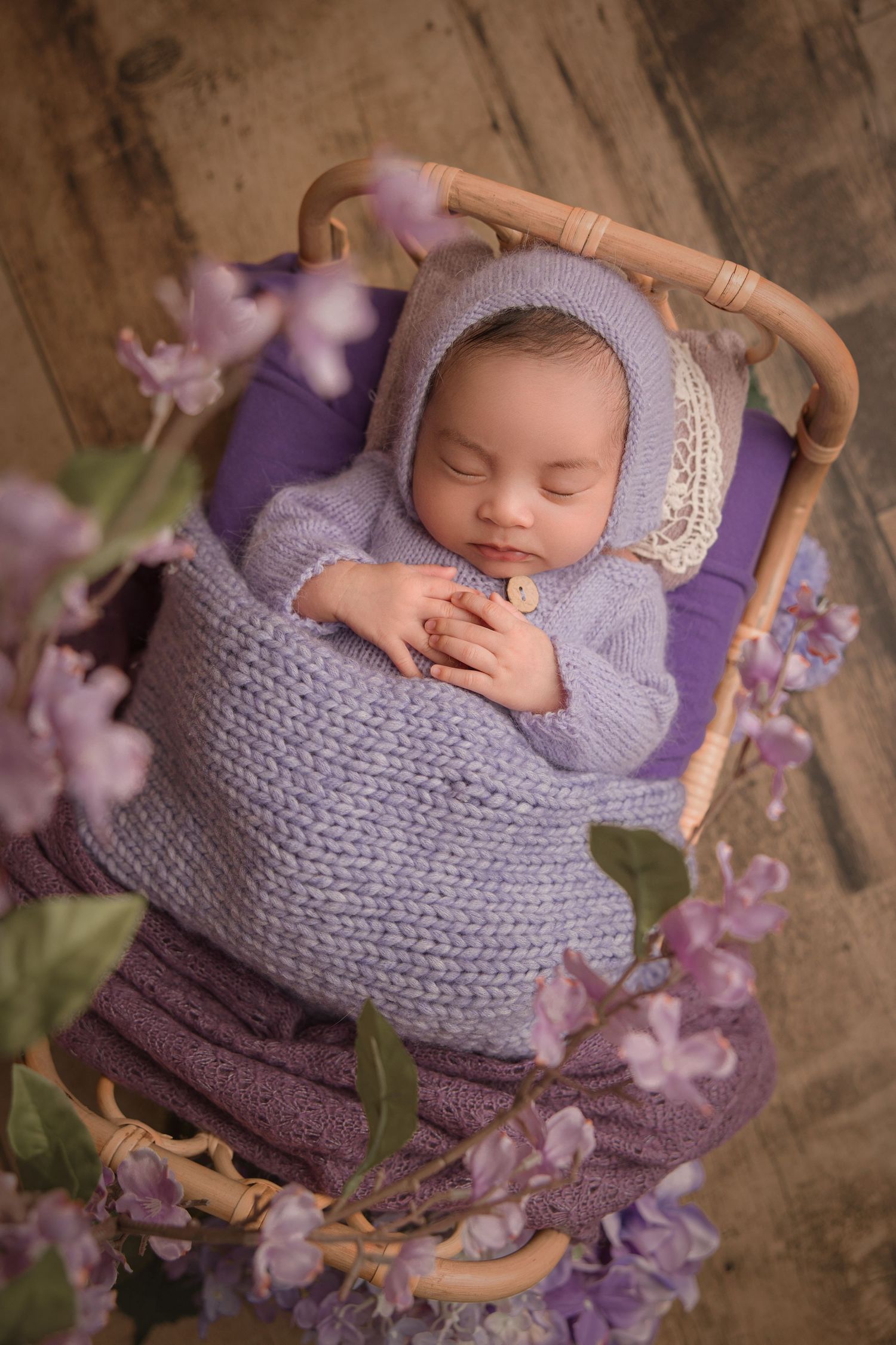 Sleeping newborn swaddled in purple and gray knit wraps nestled in wooden basket with delicate purple flowers.